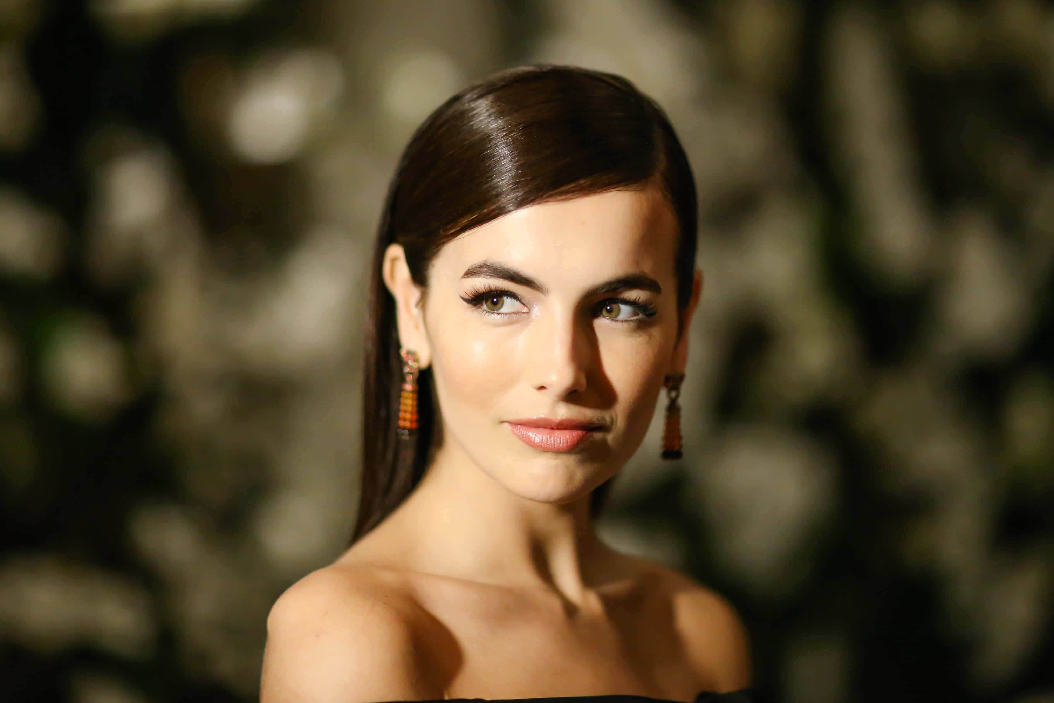 Camilla Belle 桌布 壁紙 — 一個特寫肖像，一位深色頭髮、眼睛引人注目的女性，戴著垂墜耳環，背景是模糊的自然景色。