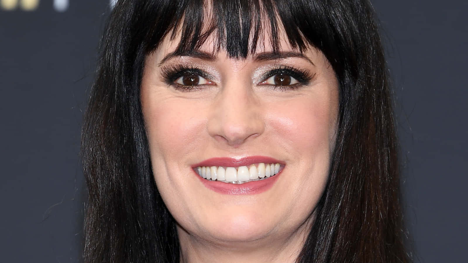 paget brewster 桌布 壁紙 — 一個特寫肖像，一位深色頭髮和劉海的女性在深灰色背景前溫暖地微笑。