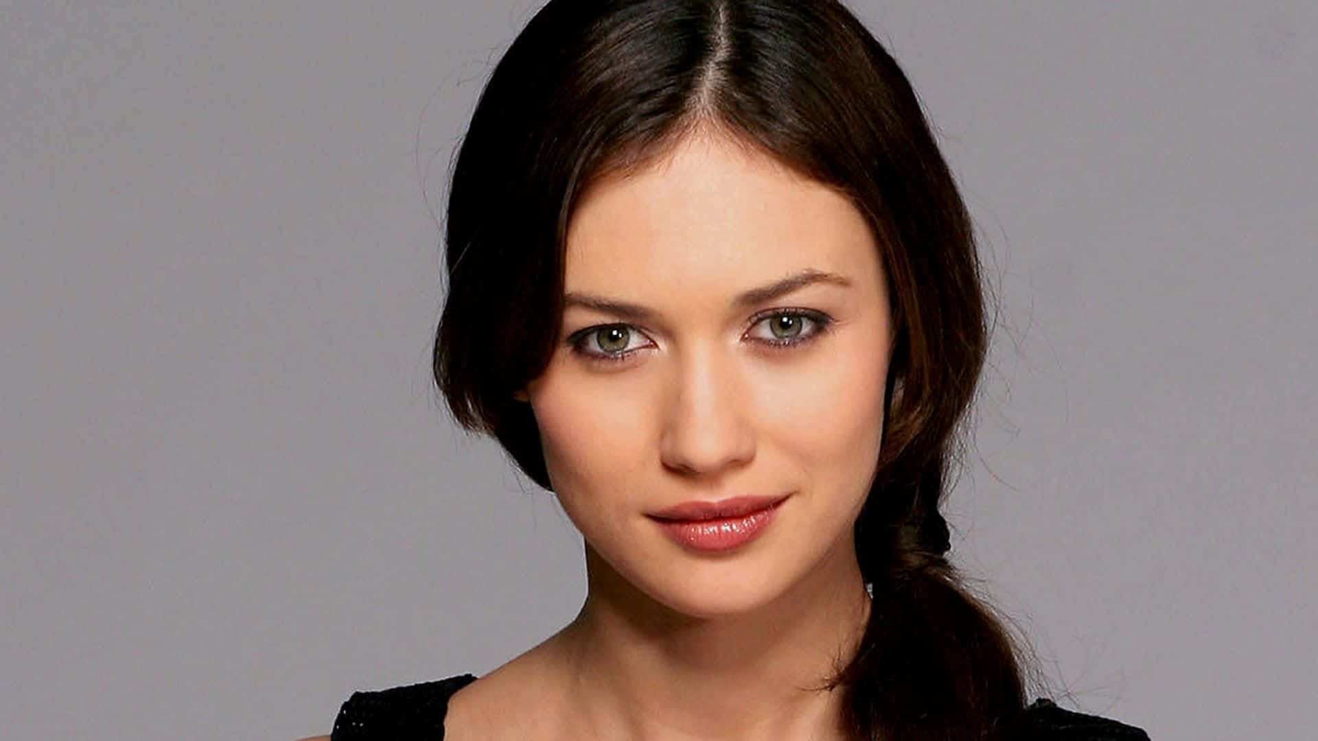 Olga Kurylenko 桌布 壁紙 — 一個特寫肖像，一位深色頭髮和綠色眼睛的女性在純灰色背景下直視鏡頭。