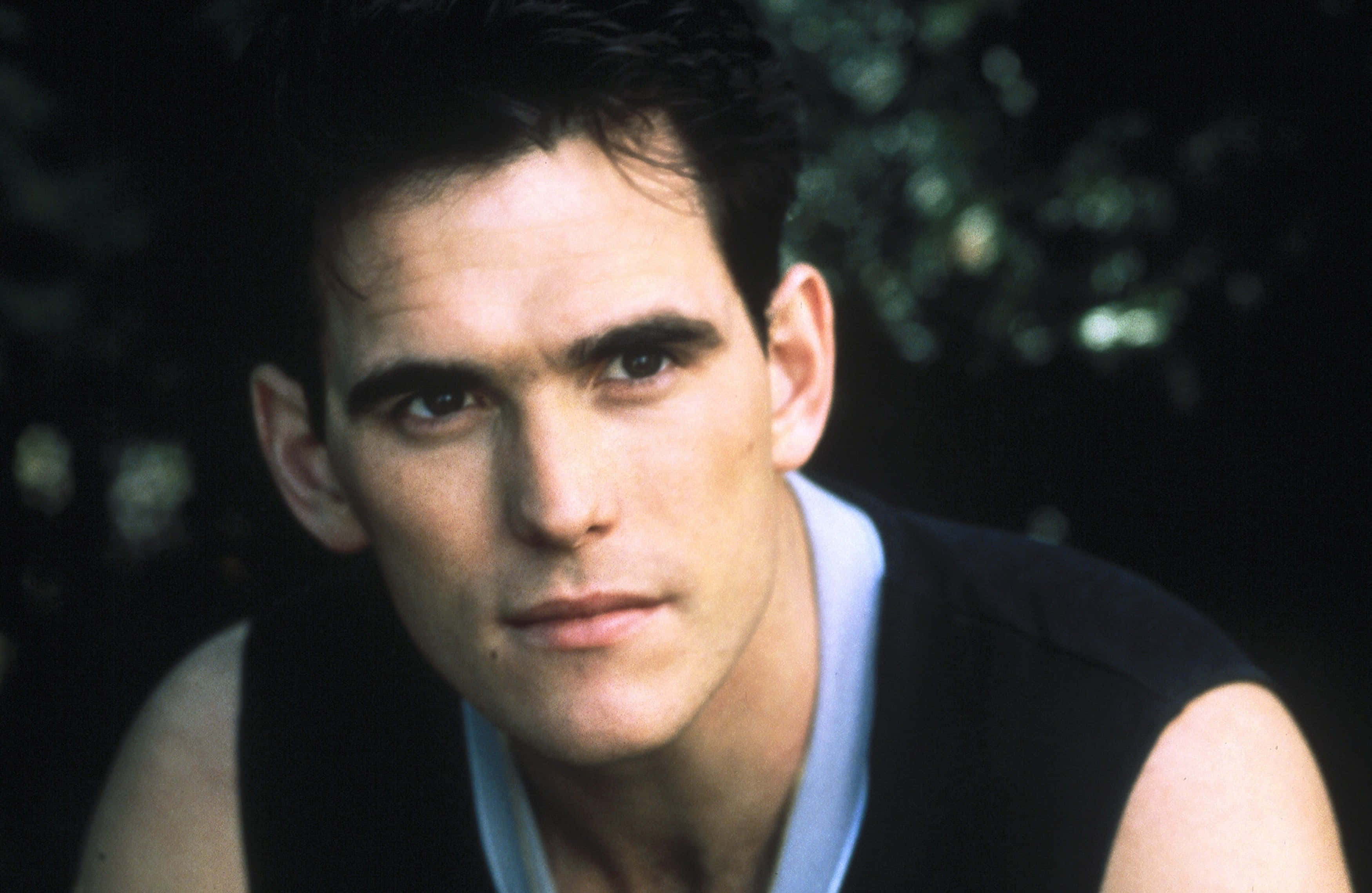 matt dillon 桌布 壁紙 — 一個特寫肖像，一個黑髮、眼神銳利的男人，穿著黑色背心，內搭淺藍色襯衫，背景是模糊的綠色樹葉。