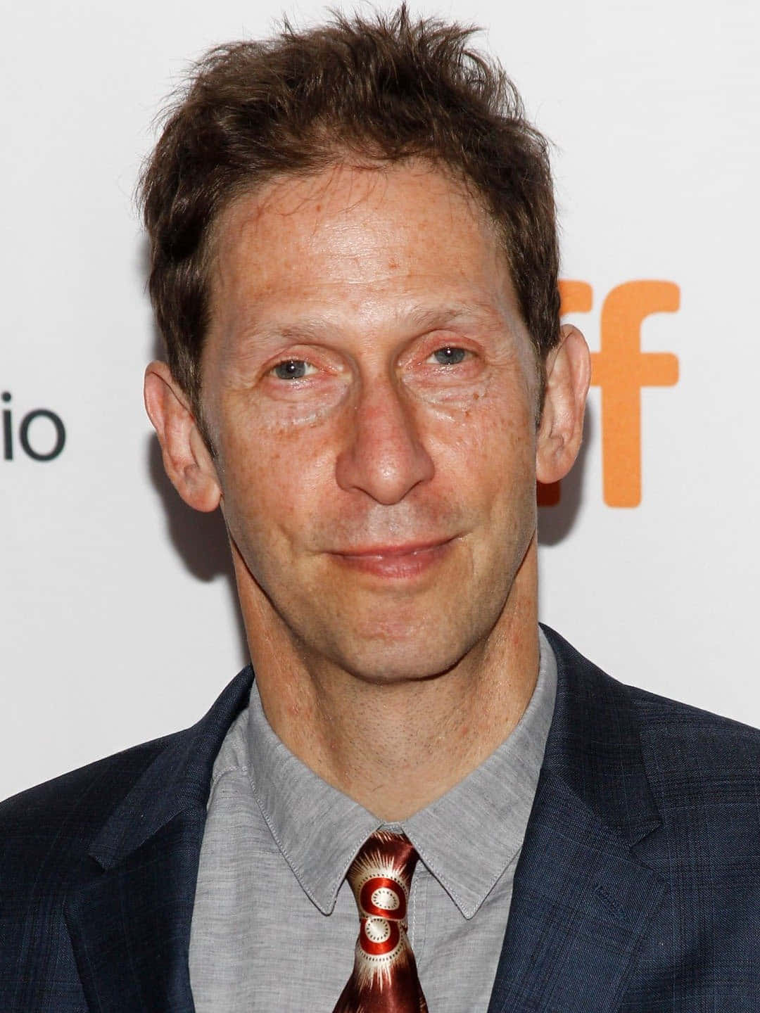 Tim Blake Nelson 桌布 壁紙 — 一個特寫肖像，一名棕色亂髮的男子穿著深色西裝和圖案領帶，背景為白色並帶有橙色字樣。