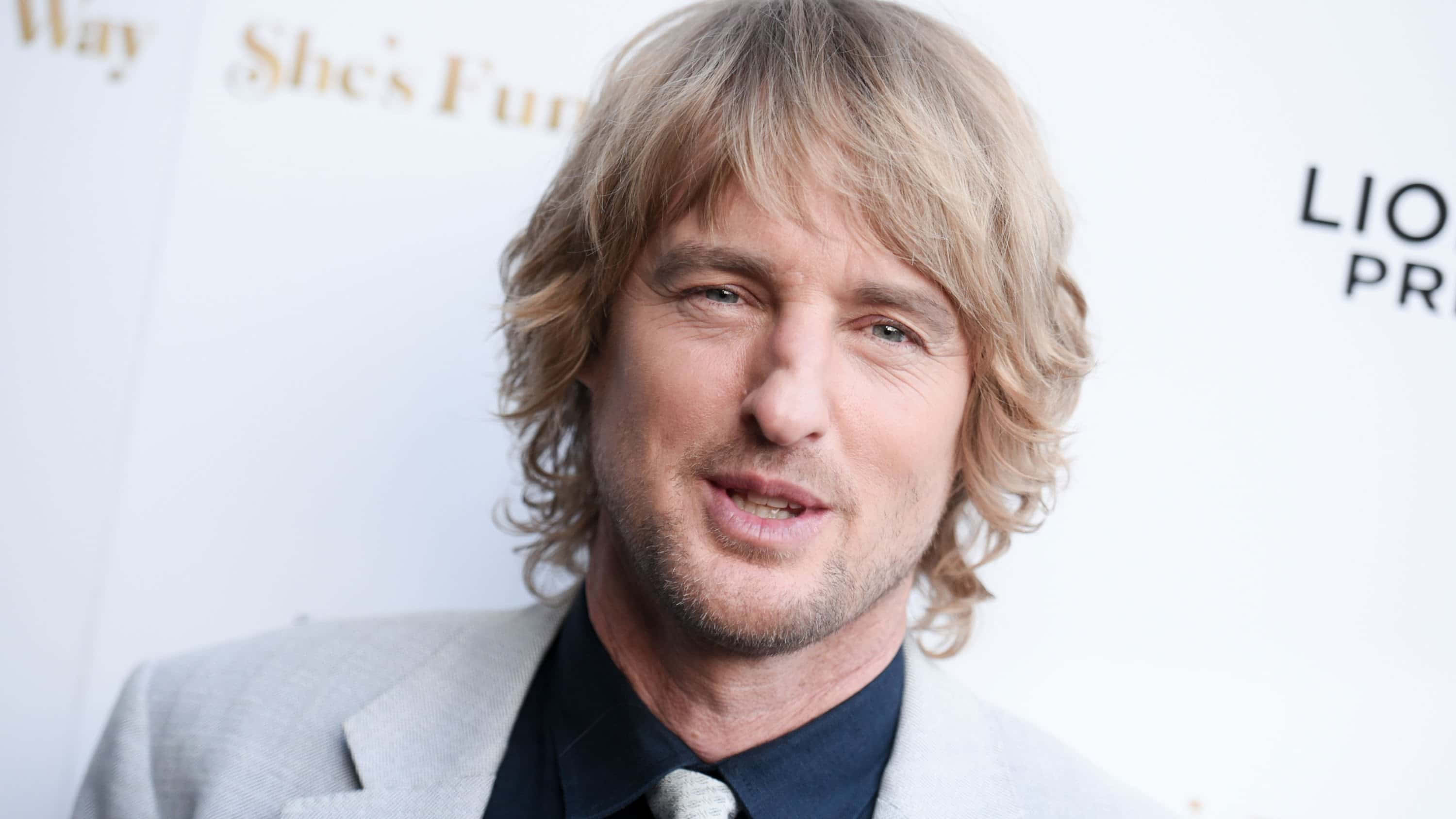 Owen Wilson 桌布 壁紙 — 一個特寫肖像，一名波浪形金髮男子穿著淺灰色西裝和深色襯衫，背景是白色活動背景。