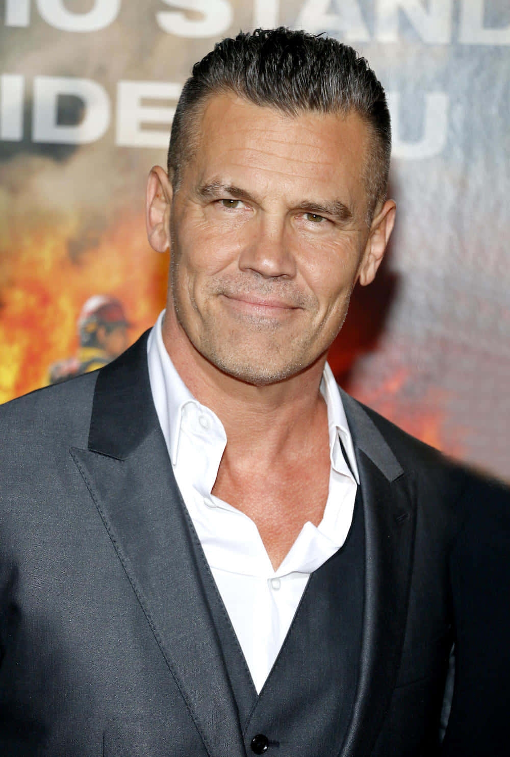 Josh Brolin 桌布 壁紙 — 一個特寫肖像，一名男子梳著深色頭髮，穿著灰色西裝和白色襯衫，背景是火熱的電影海報。