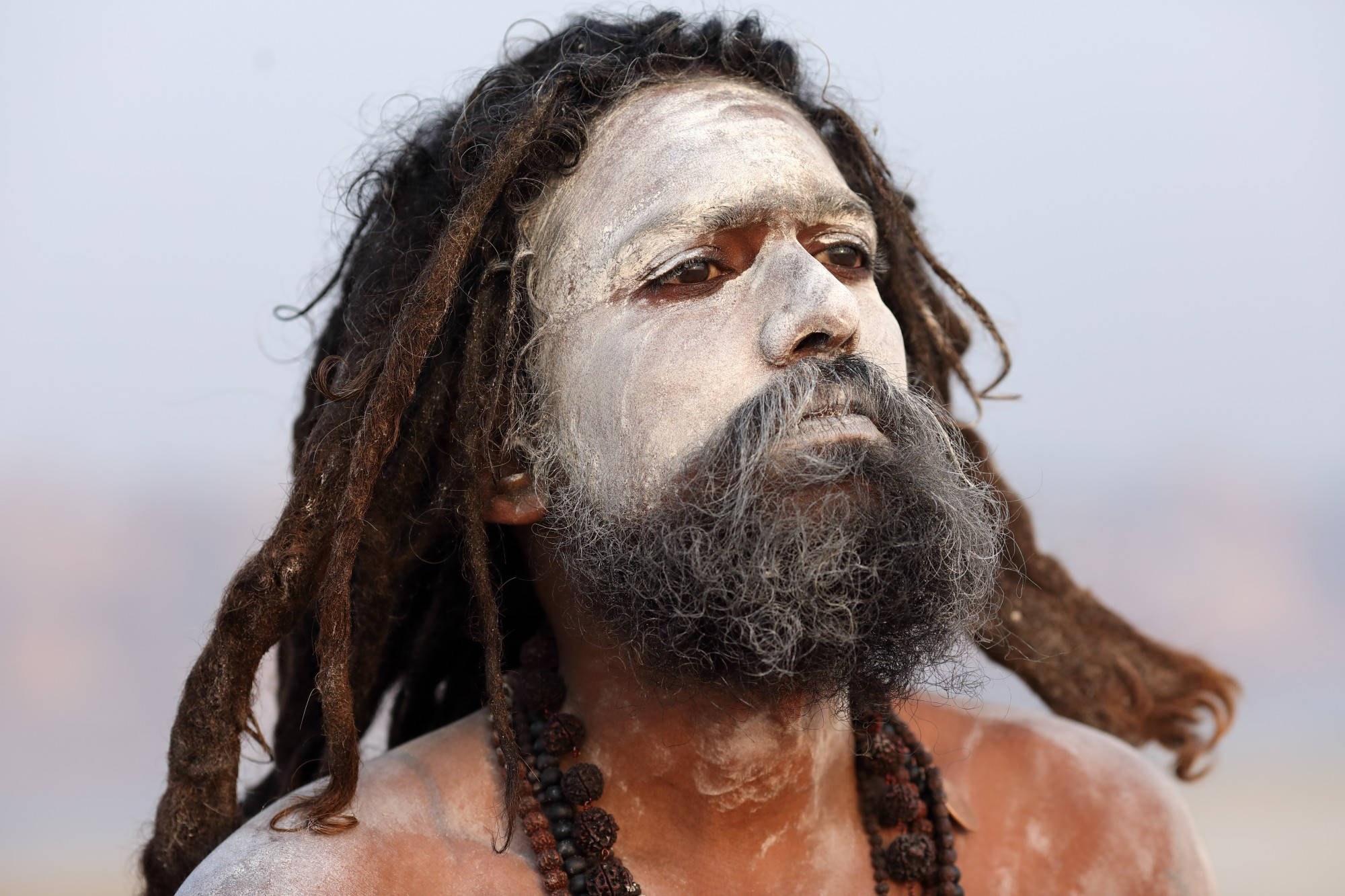 Aghori 桌布 壁紙 — 一個特寫肖像，一名留著辮子、臉上塗有白色顏料的男子，在朦朧的天空下向上凝視。