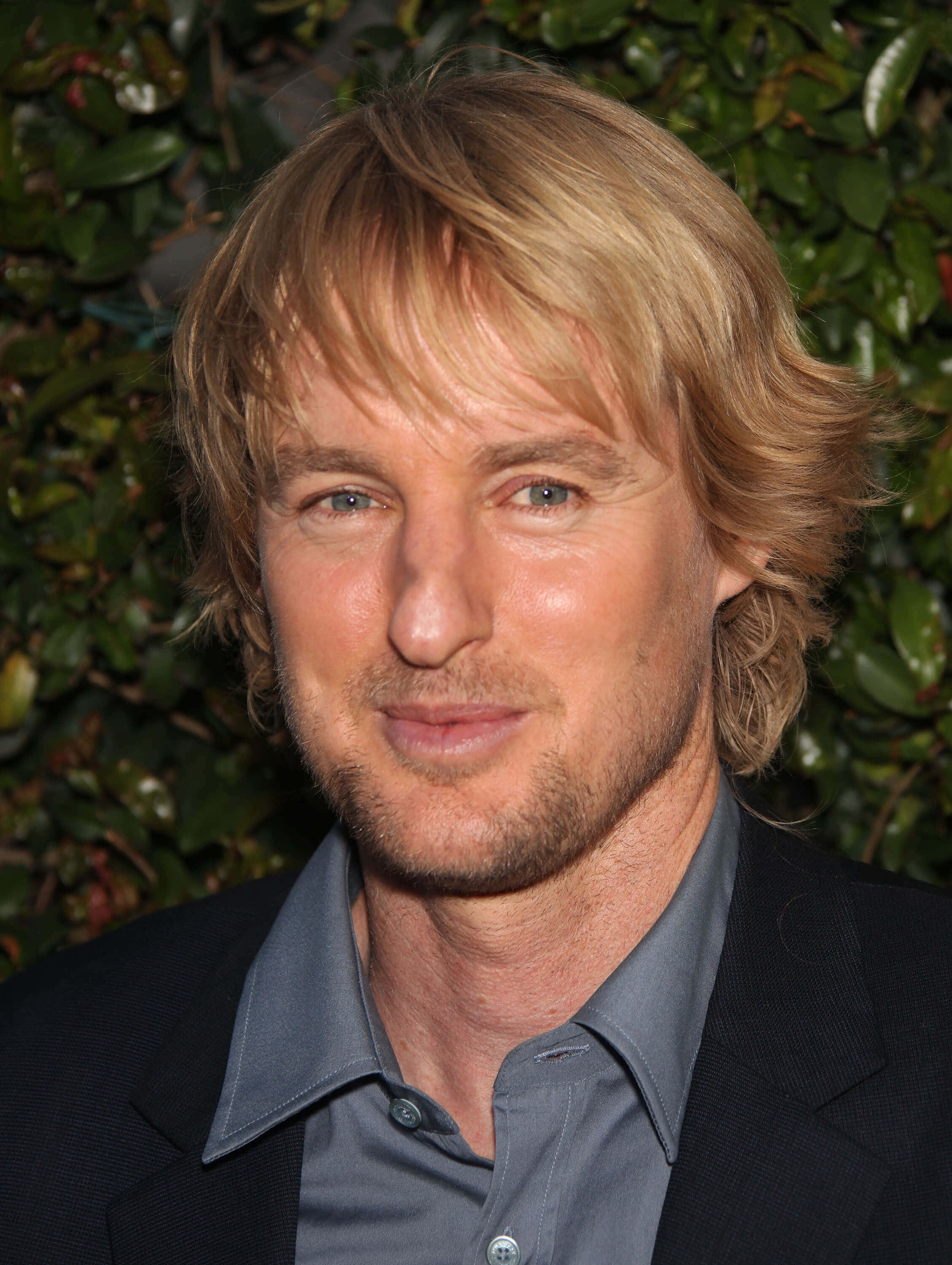 Owen Wilson 桌布 壁紙 — 一個特寫肖像，一名金髮藍眼的男士穿著深色西裝和灰色襯衫，背景是綠色樹籬。