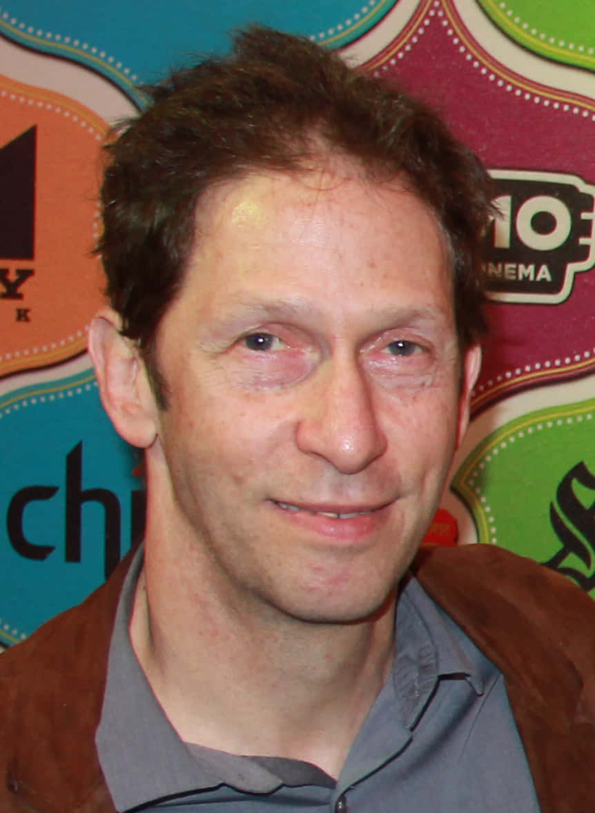 Tim Blake Nelson 桌布 壁紙 — 一個特寫肖像，一名頭髮蓬鬆的棕色頭髮男子，在色彩繽紛的圖案背景前微笑，背景上有電影院標誌。