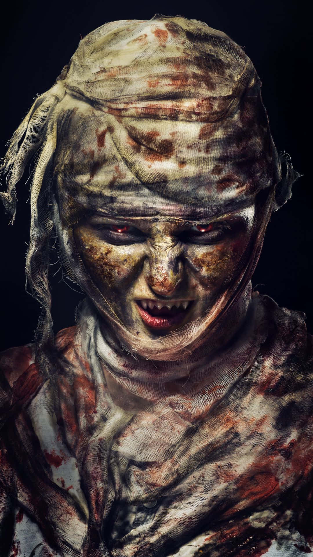 Mummy Costumes 桌布 壁紙 — 一個特寫鏡頭，展示一個怪誕的木乃伊，纏著血跡斑斑的繃帶，眼睛發出紅光，在黑色背景前兇狠地咆哮。