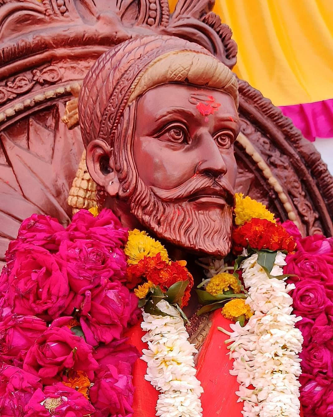 shivaji maharaj HD 桌布 壁紙 — 一個特寫鏡頭，展示了一個細節豐富的木製鬍鬚男雕像，裝飾著鮮豔的粉紅色、紅色和白色花環，背景為黃色和粉紅色。