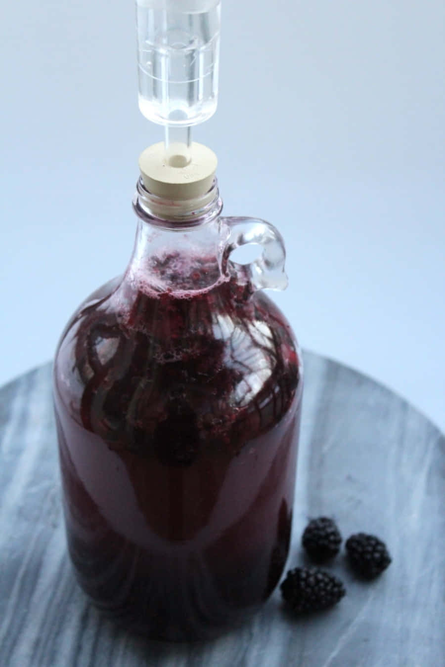 Blackberry Wine 桌布 壁紙 — 一個玻璃壺裝滿深紫色黑莓液體和發酵氣閥，放在灰色木質表面上，附近有新鮮黑莓。