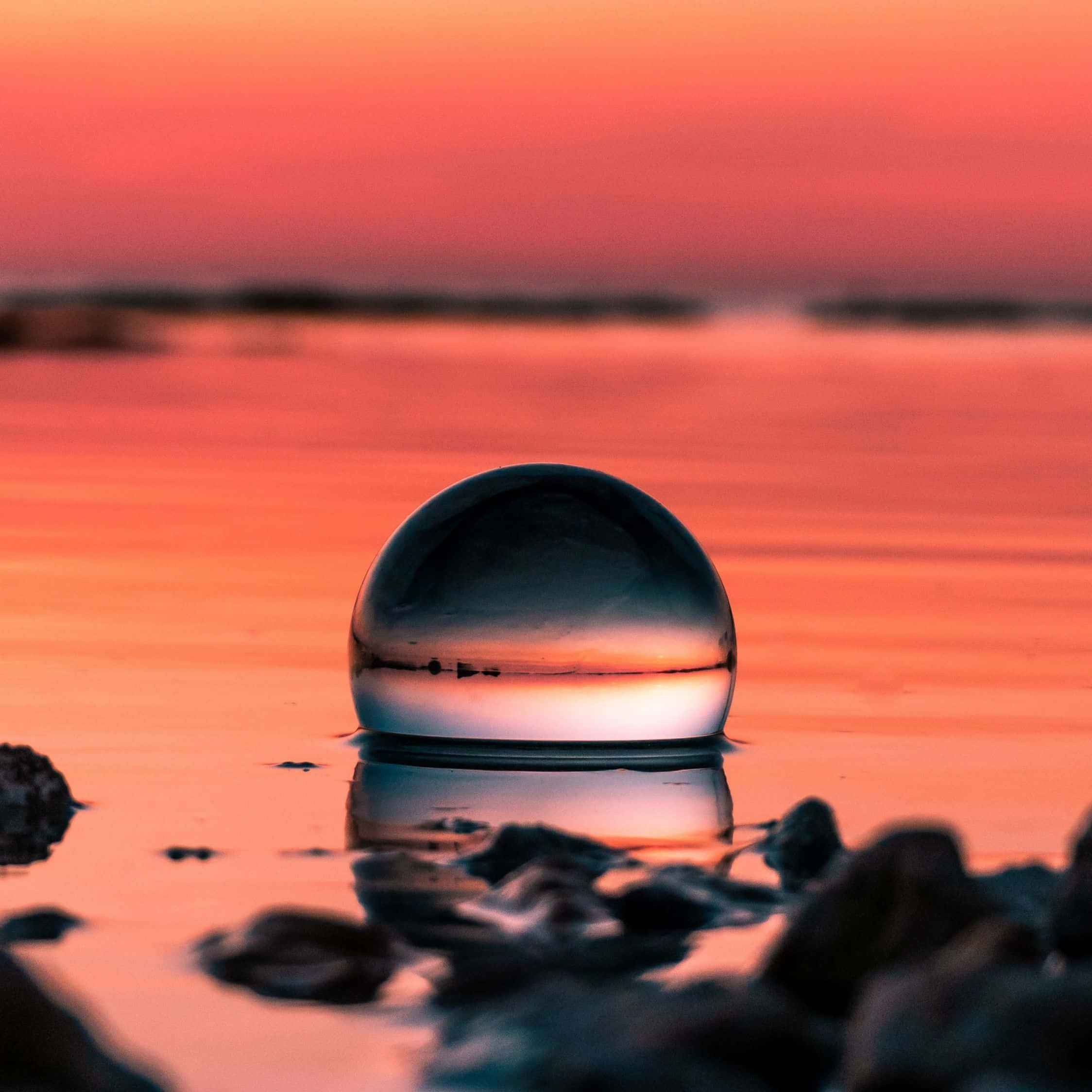 Crystal Ball 桌布 壁紙 — 一個玻璃球在日落時漂浮在水面上，反射出火熱的橙色和粉紅色天空，前景有模糊的岩石。