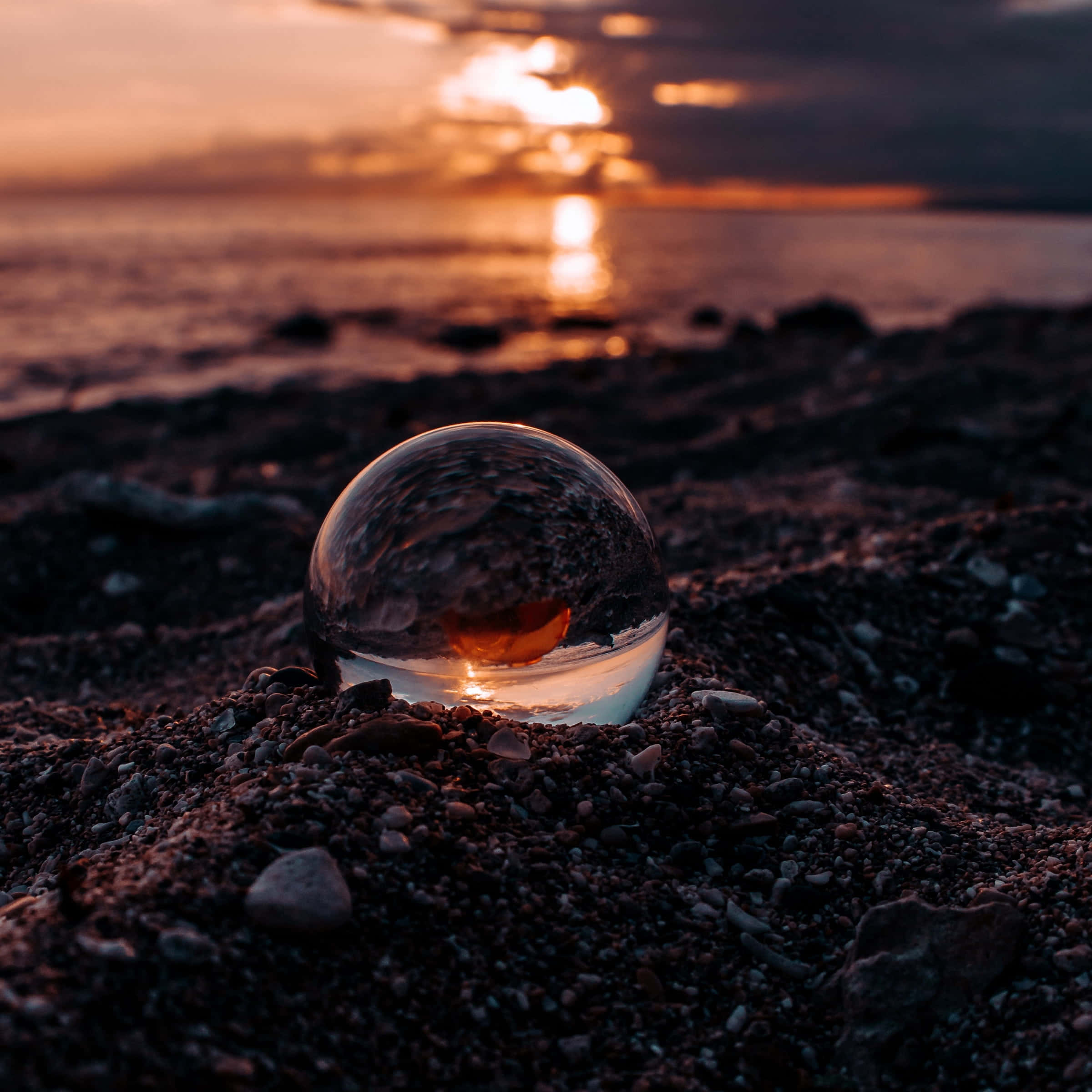 Blissful 桌布 壁紙 — 一個玻璃球體擱在卵石海灘上，反射出海洋上空熾熱的日落。