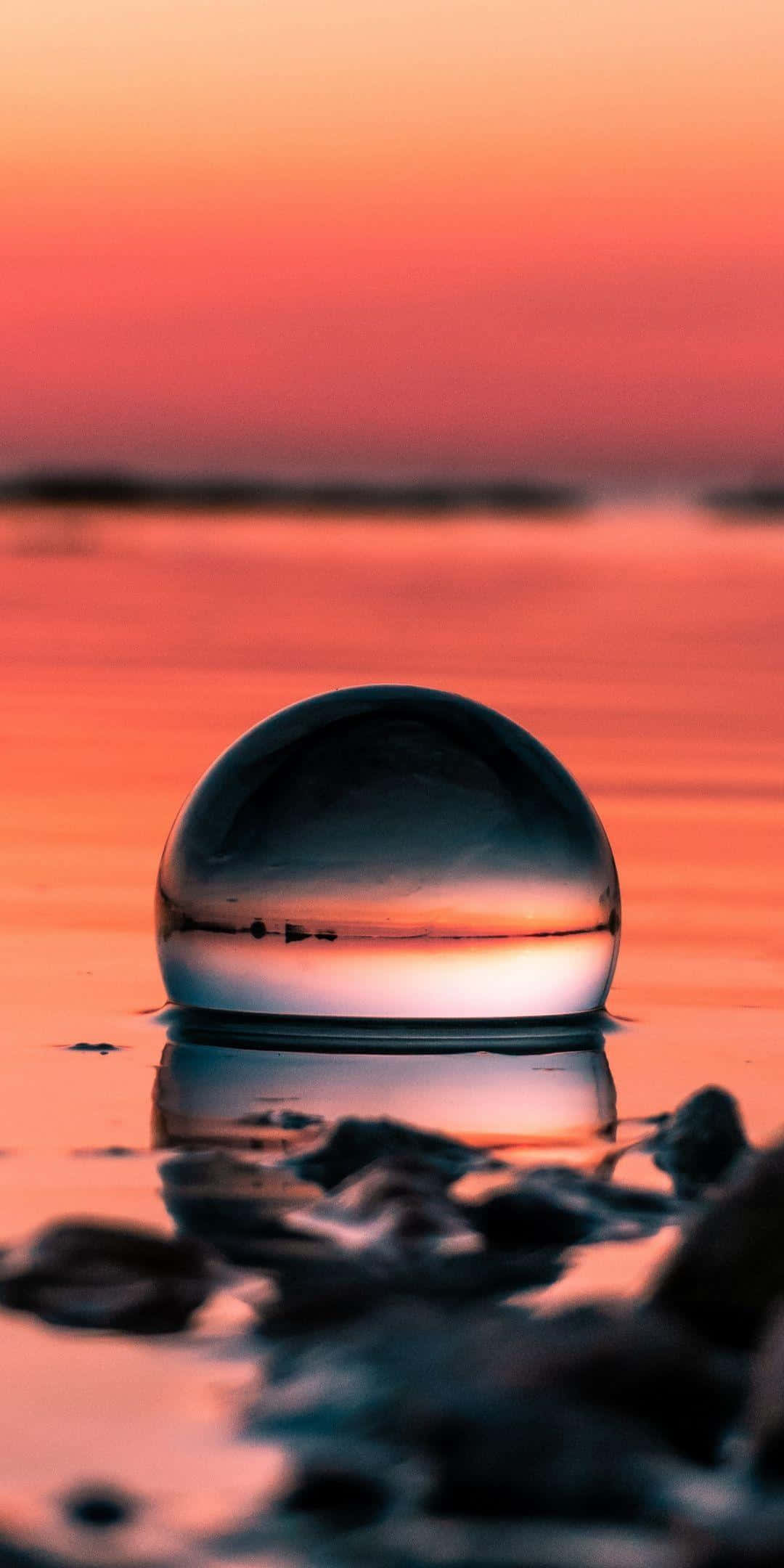 Sunset Reflection 桌布 壁紙 — 一個玻璃球體漂浮在水面上，反射出火熱的橙色日落天空和遠方地平線。