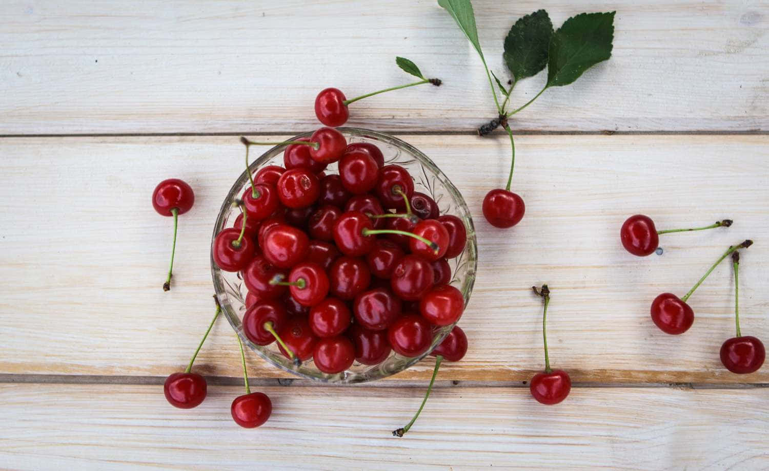 Red Cherries 桌布 壁紙 — 一個玻璃碗裝滿了光澤的紅色櫻桃，放在淺色木質表面上，周圍散落著水果和綠葉。