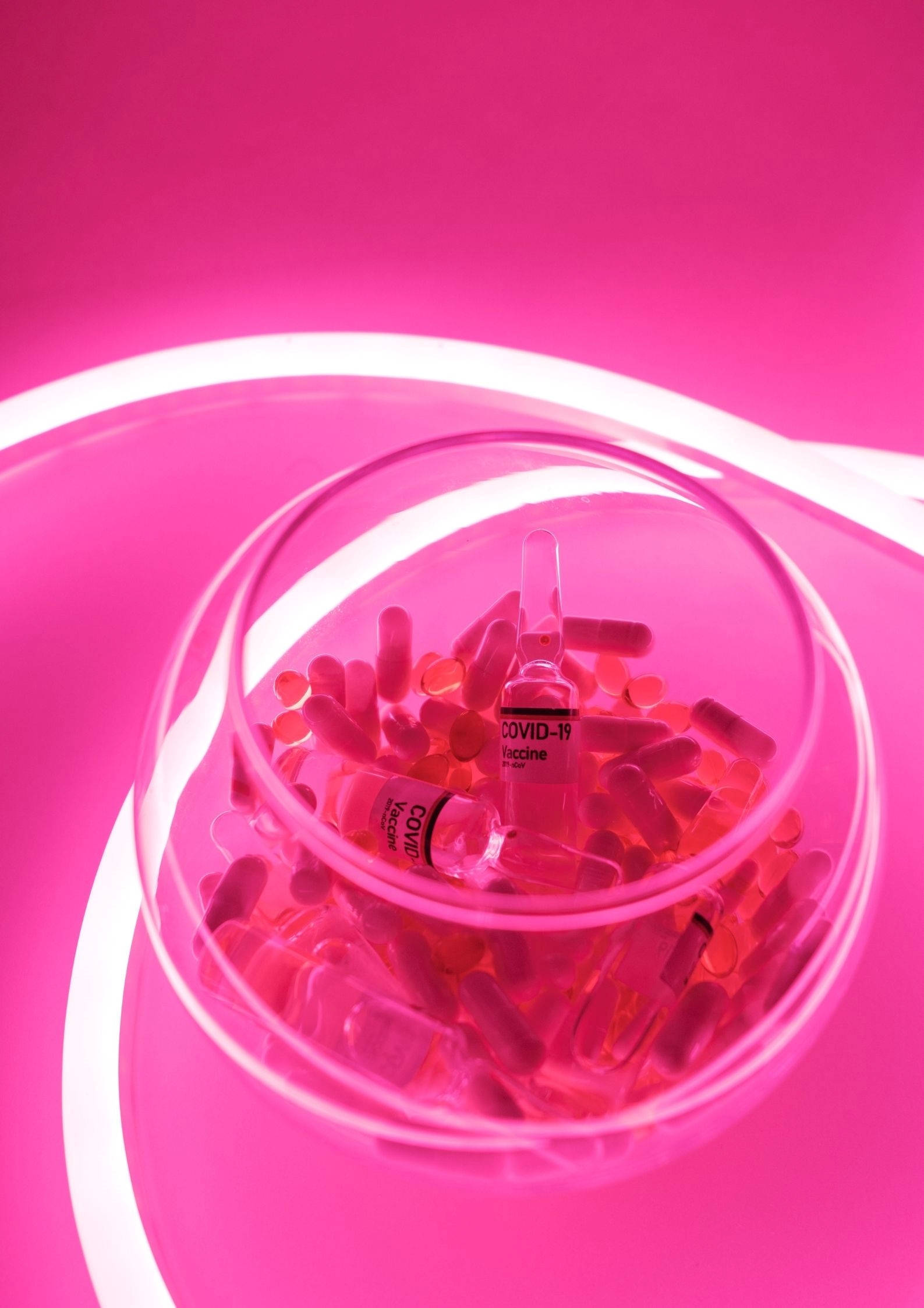可愛 桌布 壁紙 — 一個玻璃碗裝滿粉紅色的COVID-19疫苗小瓶和膠囊，放在帶有白色霓虹曲線的發光粉紅色表面上。