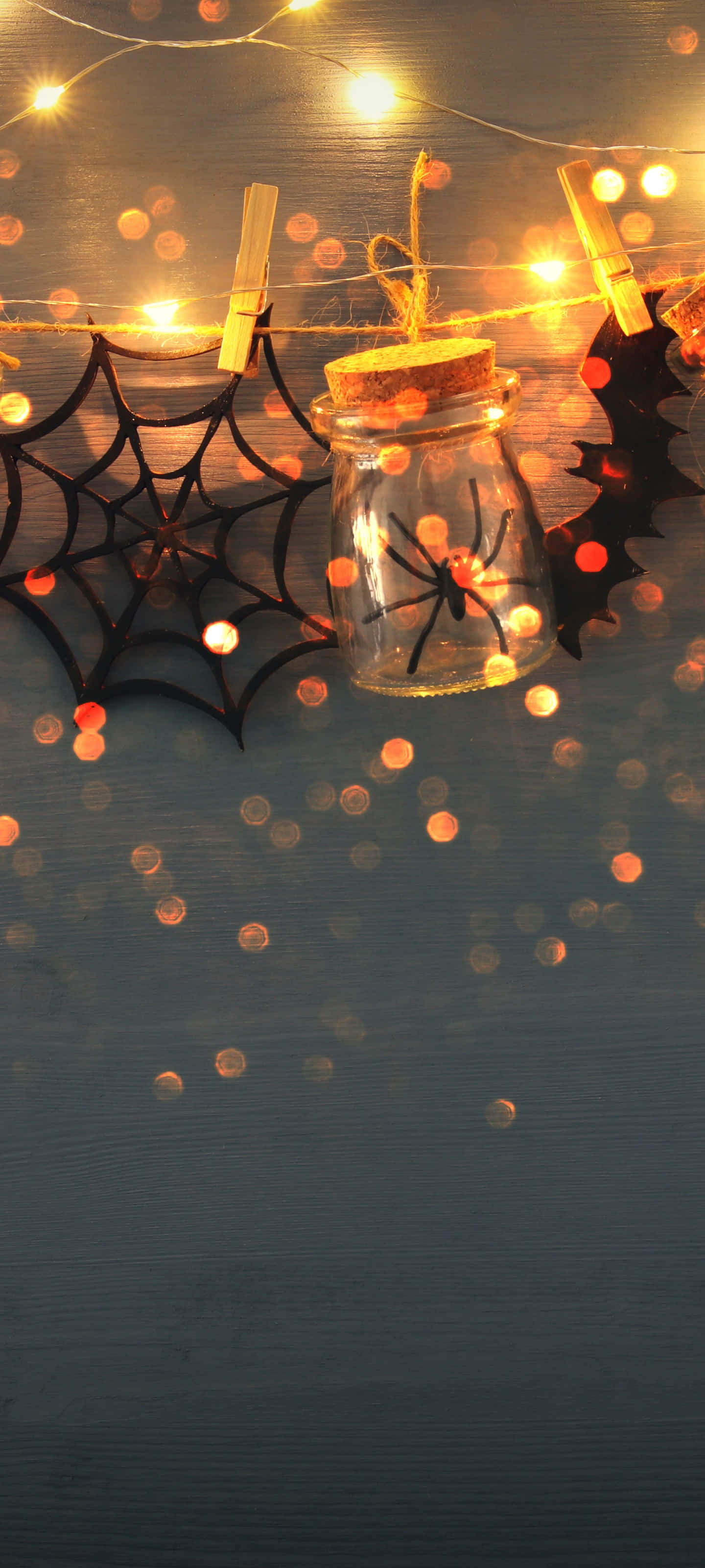 Halloween Phone 桌布 壁紙 — 一個玻璃罐內有一隻黑色蜘蛛，放在深色木質表面上，旁邊有蜘蛛網和發光的燈串。