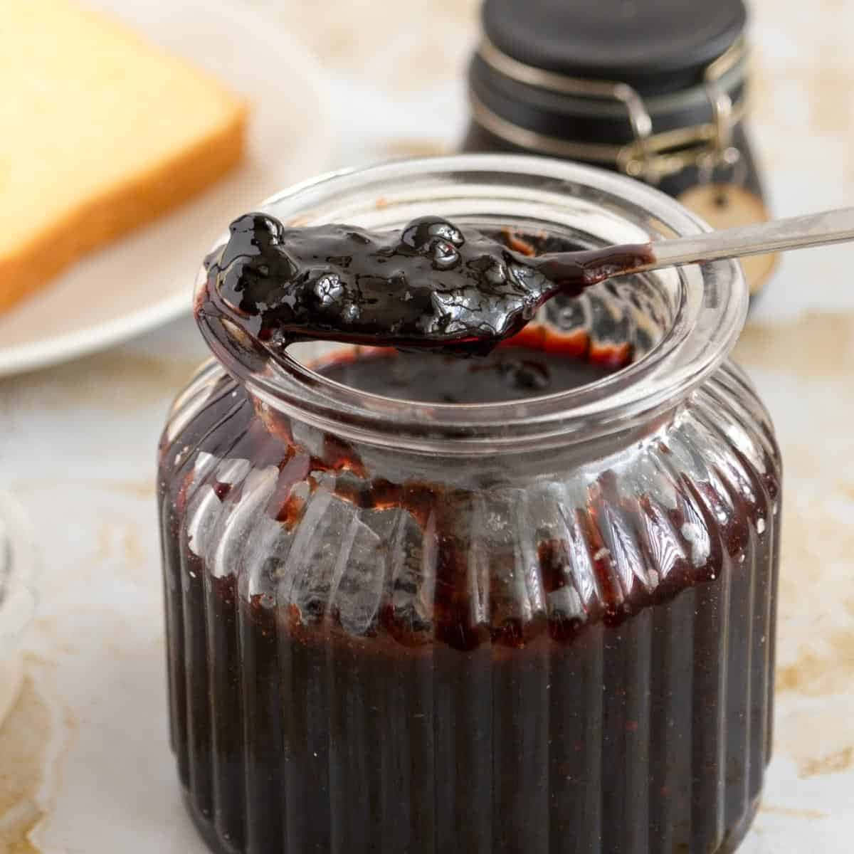 Blueberry Jam 桌布 壁紙 — 一個玻璃罐裝滿了光澤的深色莓果醬，湯匙舀起一勺，呈現濃稠質地，旁邊放著一片麵包。