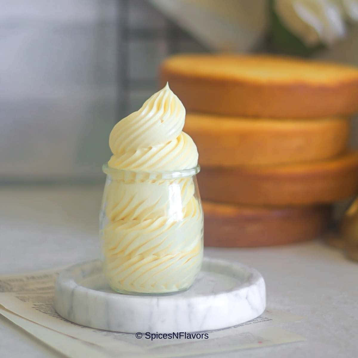 Buttercream 桌布 壁紙 — 一個玻璃罐裝滿了奶油香草牛油忌廉，放在大理石杯墊上，背景有模糊的蛋糕層。