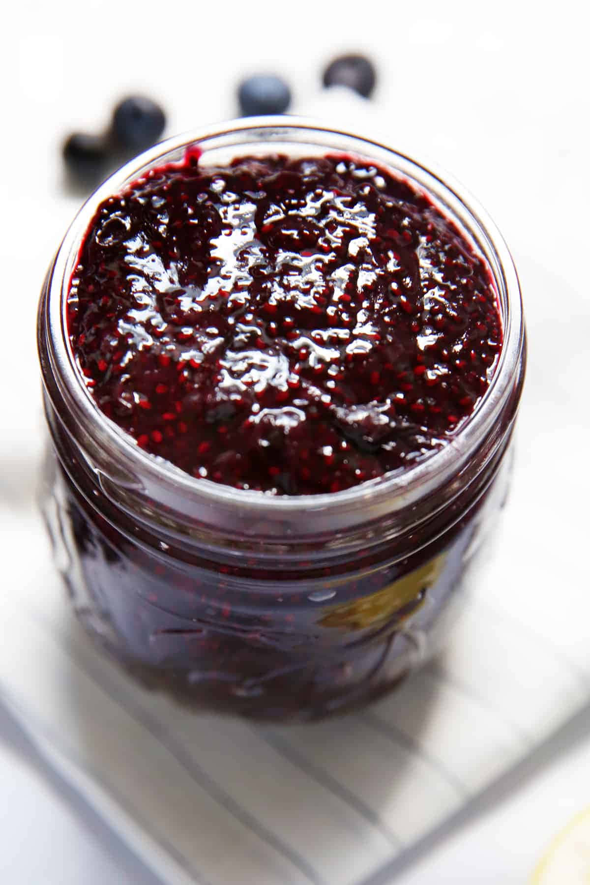 Blueberry Jam 桌布 壁紙 — 一個玻璃罐裝滿光澤深藍色藍莓醬，放在白色條紋表面上，附近散落著新鮮藍莓。