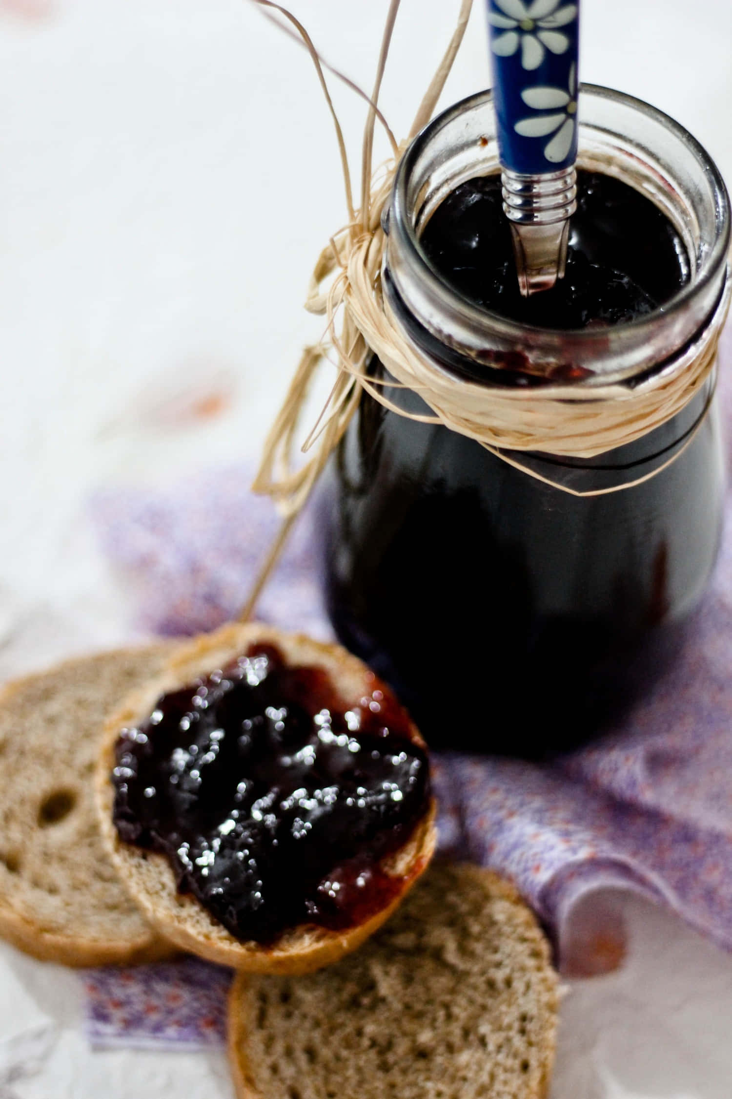 Blackberry Jam 桌布 壁紙 — 一個玻璃罐裝的深色莓果醬，配上一把花卉圖案的勺子，放在圖案布上的鄉村麵包片旁邊。