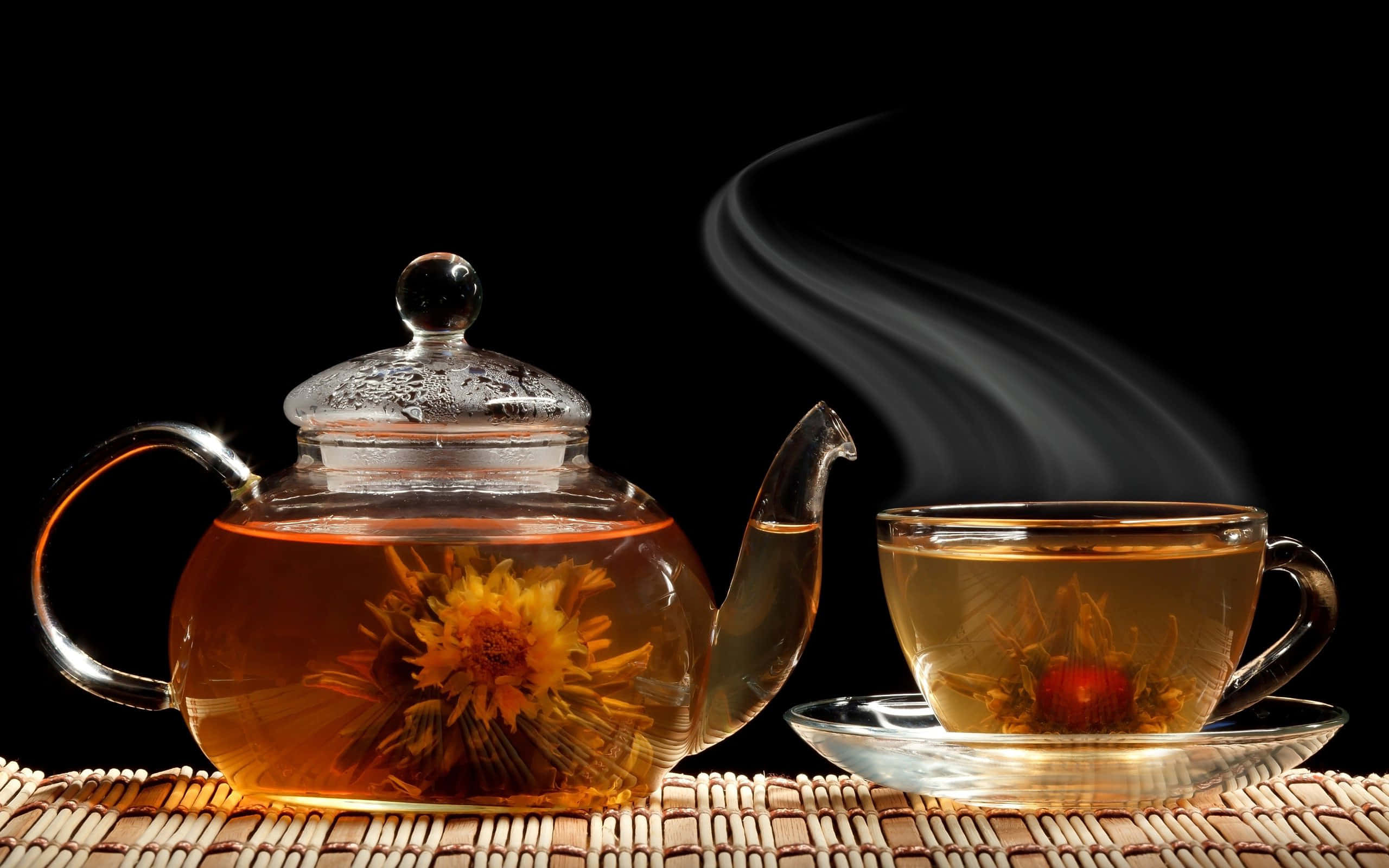 Floral Tea 桌布 壁紙 — 一個玻璃茶壺和杯子，裝滿冒著熱氣的花草茶和盛開的花朵，放在黑色背景前的編織墊上。