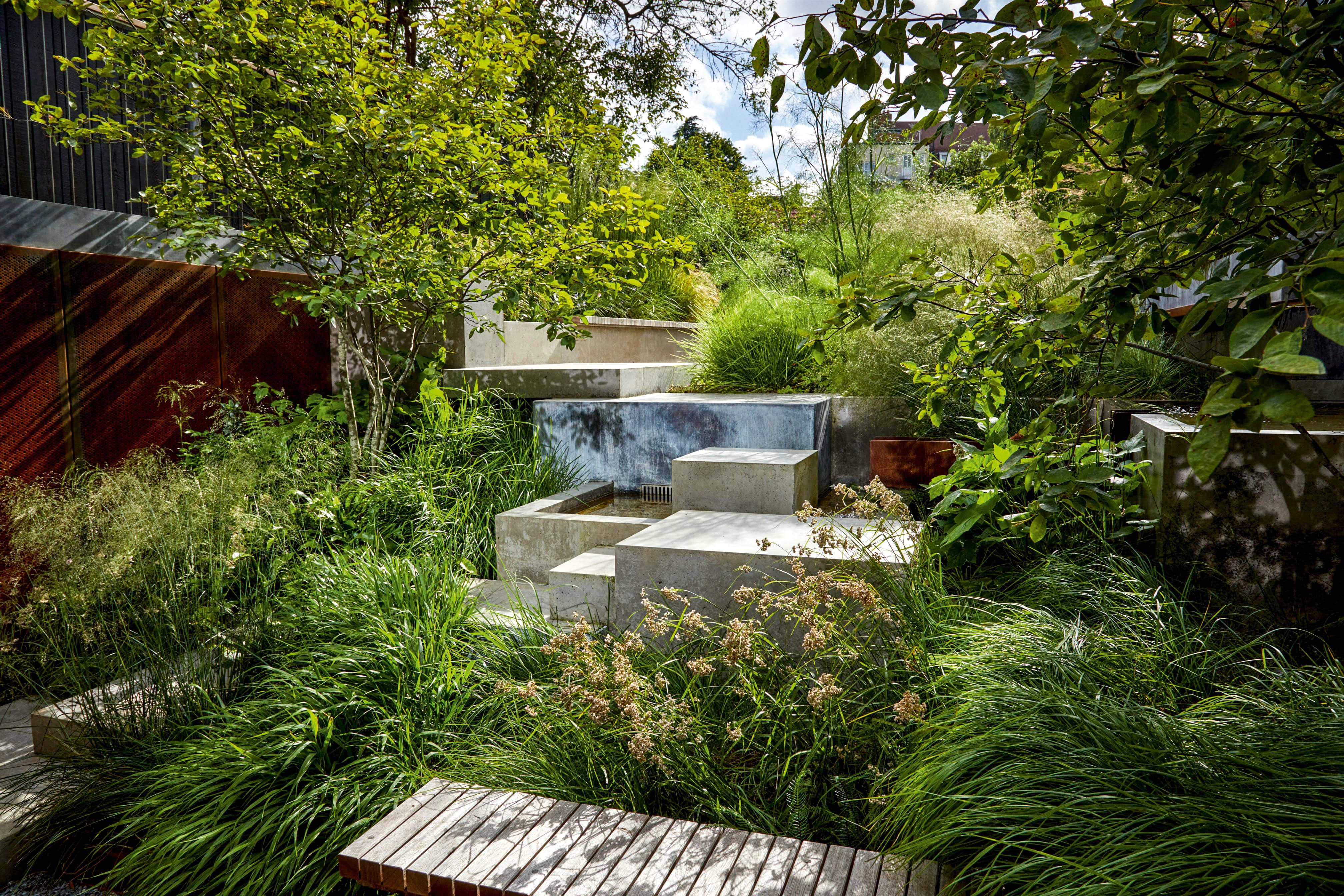 Garden Design 桌布 壁紙 — 一個現代混凝土水景，坐落在茂密的綠色花園中，有高大的草和枝葉繁茂的樹木，在明亮的天空下。