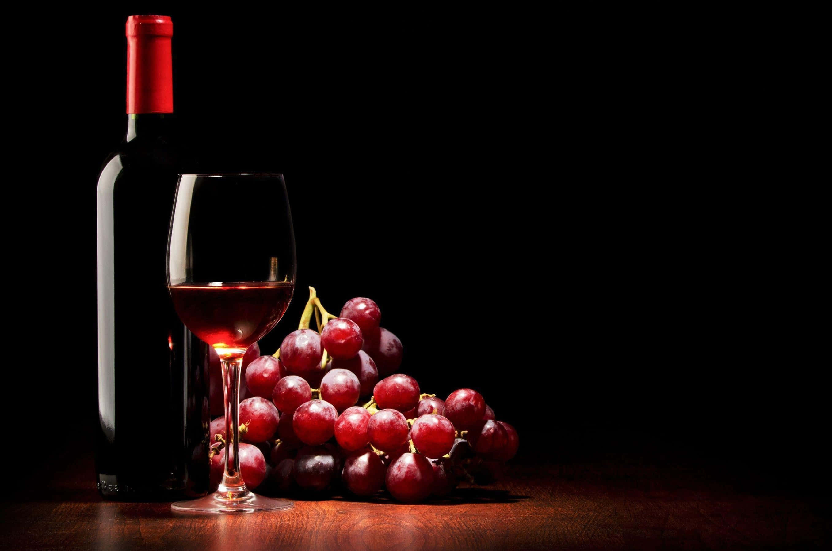 Red Wine Glass 桌布 壁紙 — 一個瓶玻璃和一串紅葡萄在黑色背景下的木質表面上被照亮。