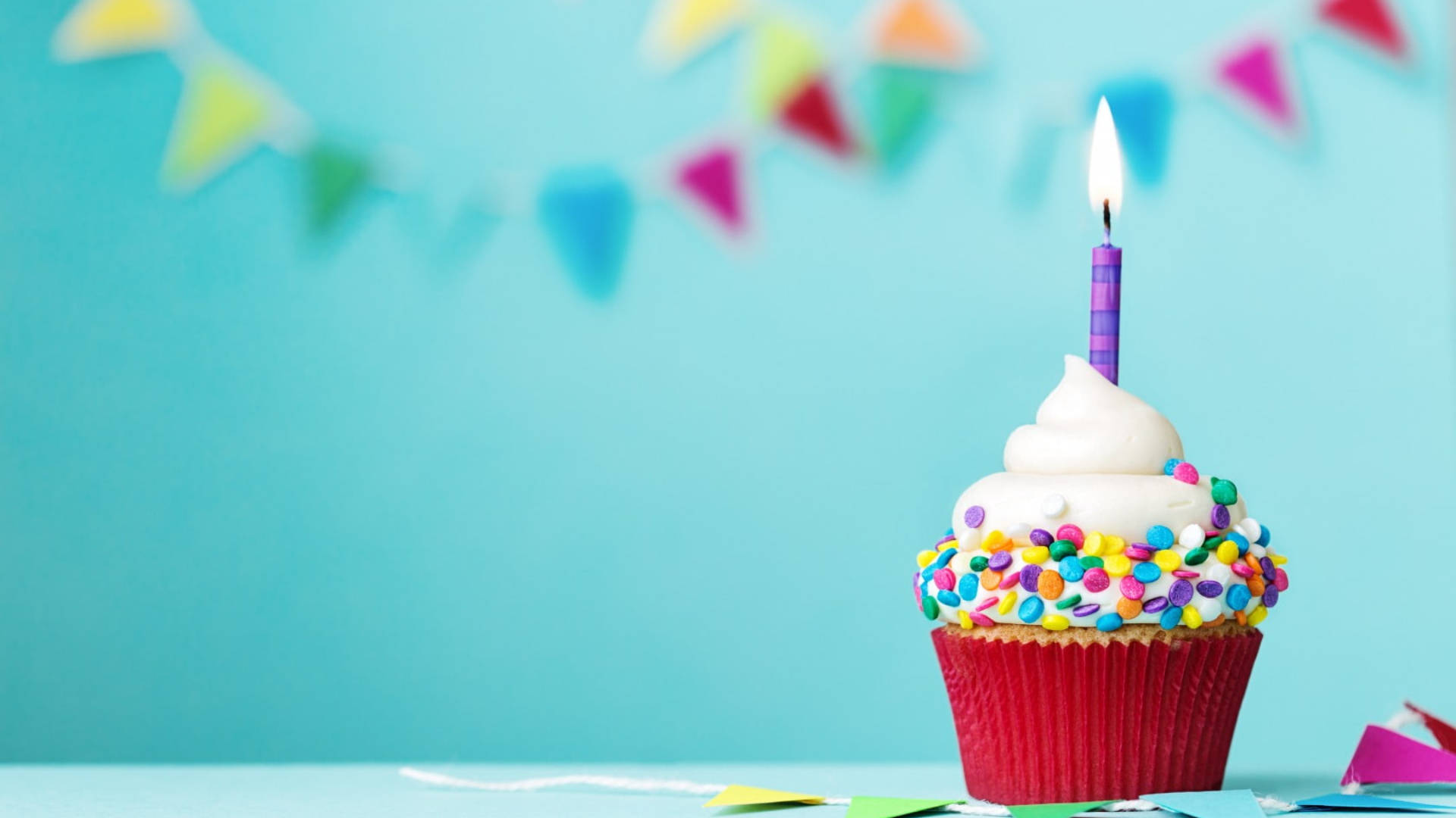 Birthday Party 桌布 壁紙 — 一個生日杯子蛋糕，上面有白色糖霜和彩色糖粒，頂部插著一根點燃的紫色蠟燭，背景是藍綠色，帶有模糊的派對旗幟。