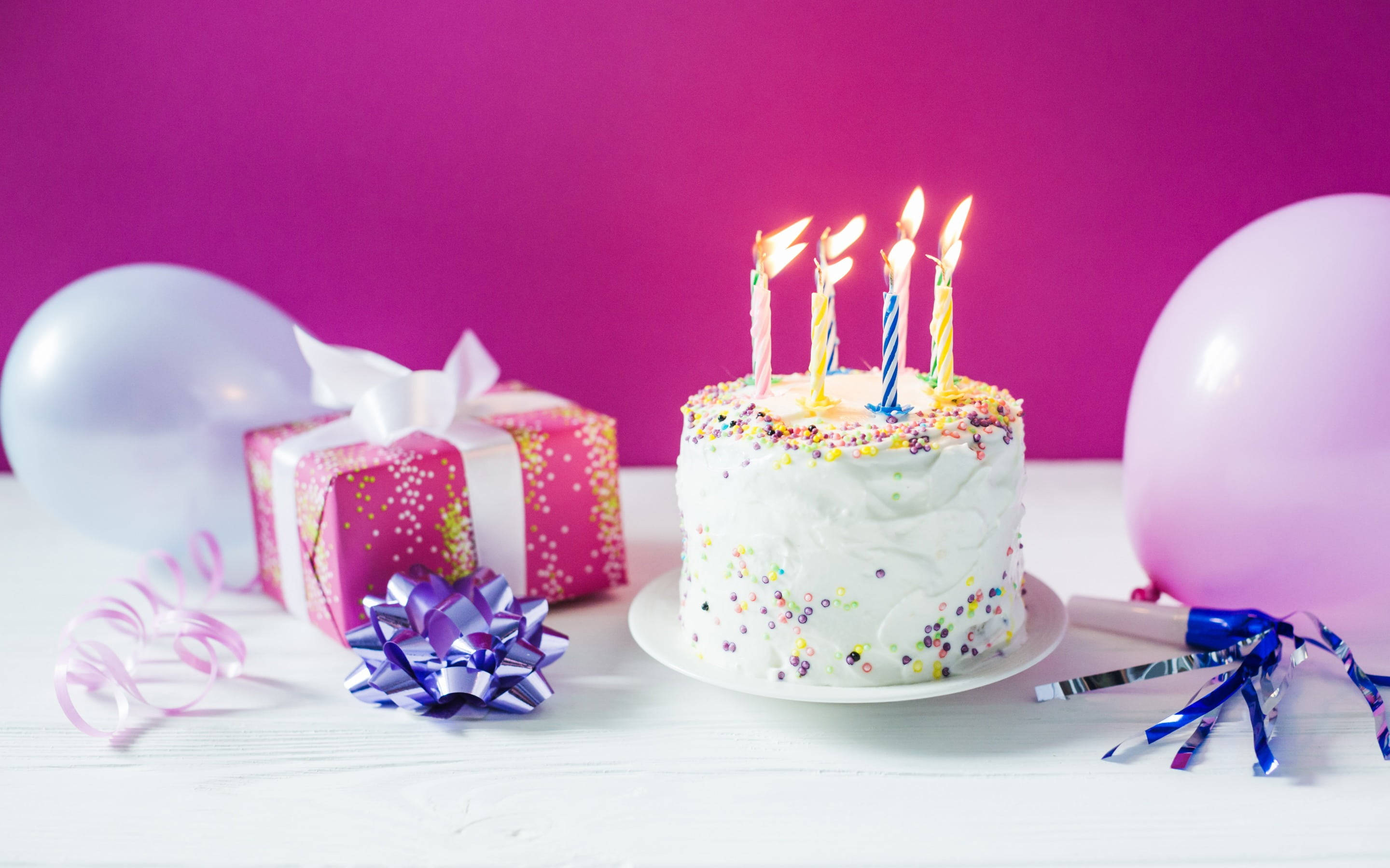 生日蛋糕 桌布 壁紙 — 一個生日蛋糕，上面有燃燒的蠟燭和彩色糖粒，旁邊放著禮物、氣球和彩帶，放在白色桌子上，背景是鮮豔的粉紅色。