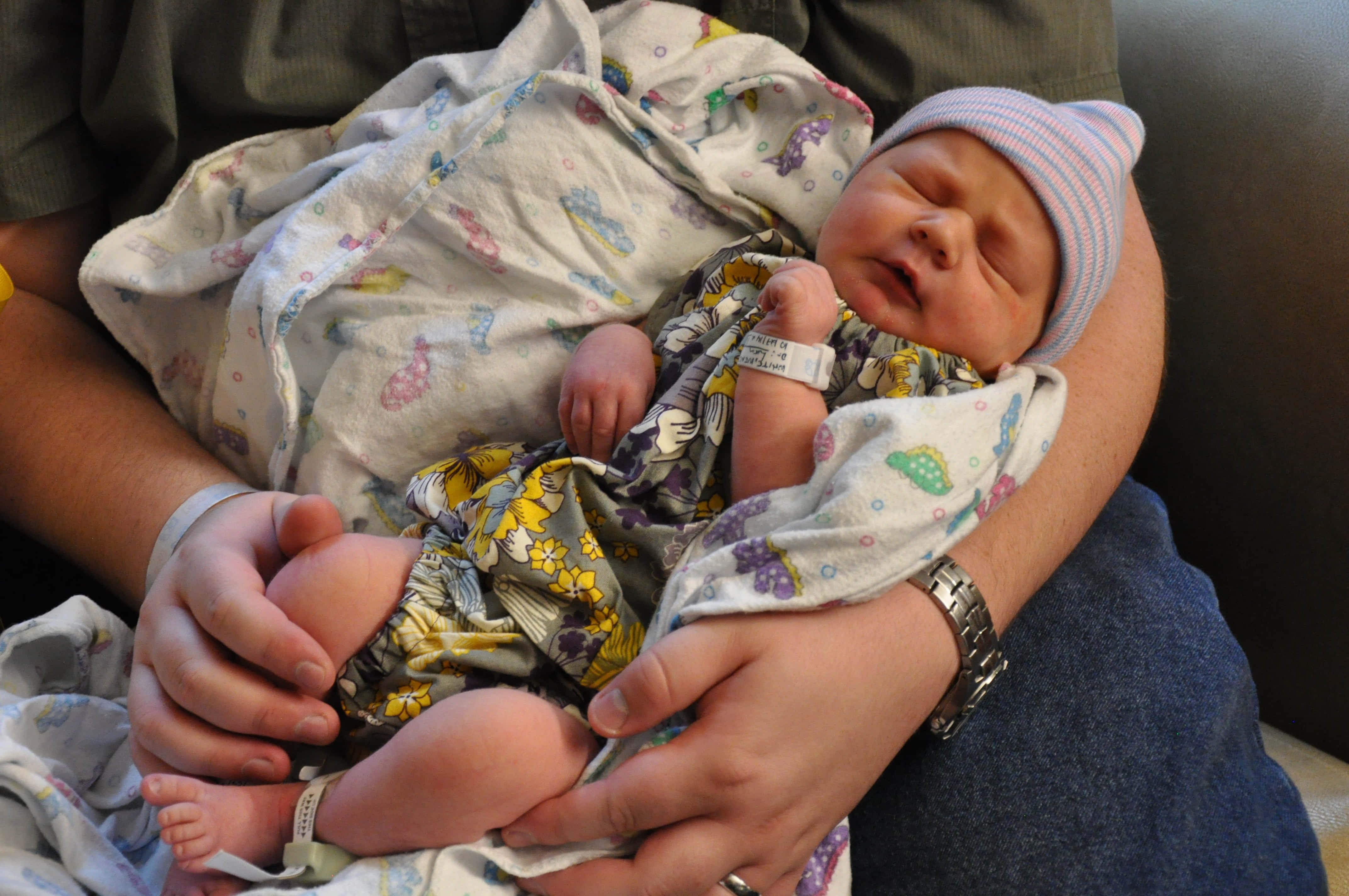 Newborn Baby 桌布 壁紙 — 一個用圖案毛毯包裹的新生嬰兒，戴著條紋帽子和醫院手帶，在成人懷中安詳地睡覺。