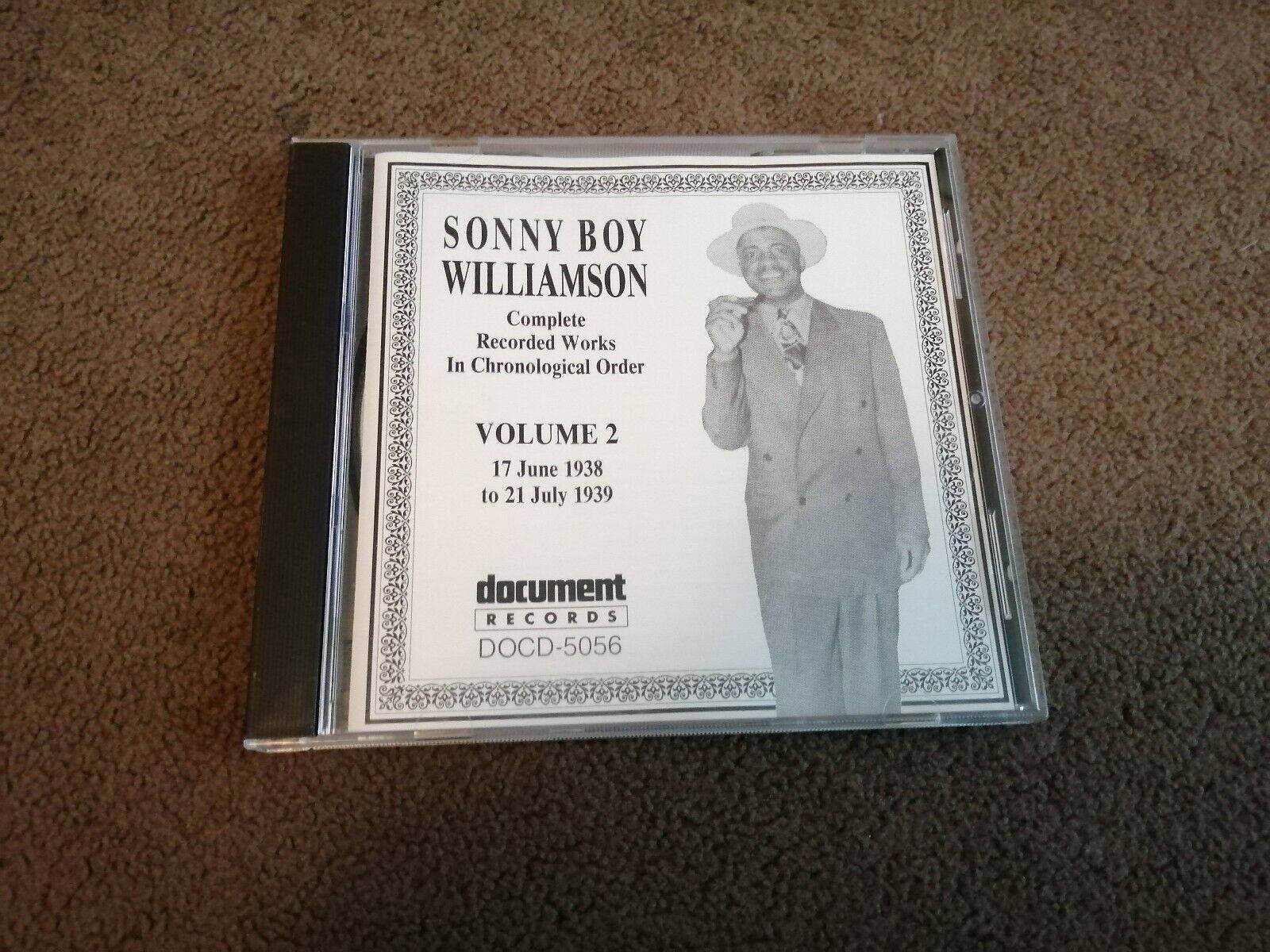 Sonny Boy Williamson II《Complete Document Records》 壁紙 — 一個用於Sonny Boy Williamson音樂的復古CD盒放在棕色地毯上，上面有藝術家穿著西裝和帽子的黑白照片。