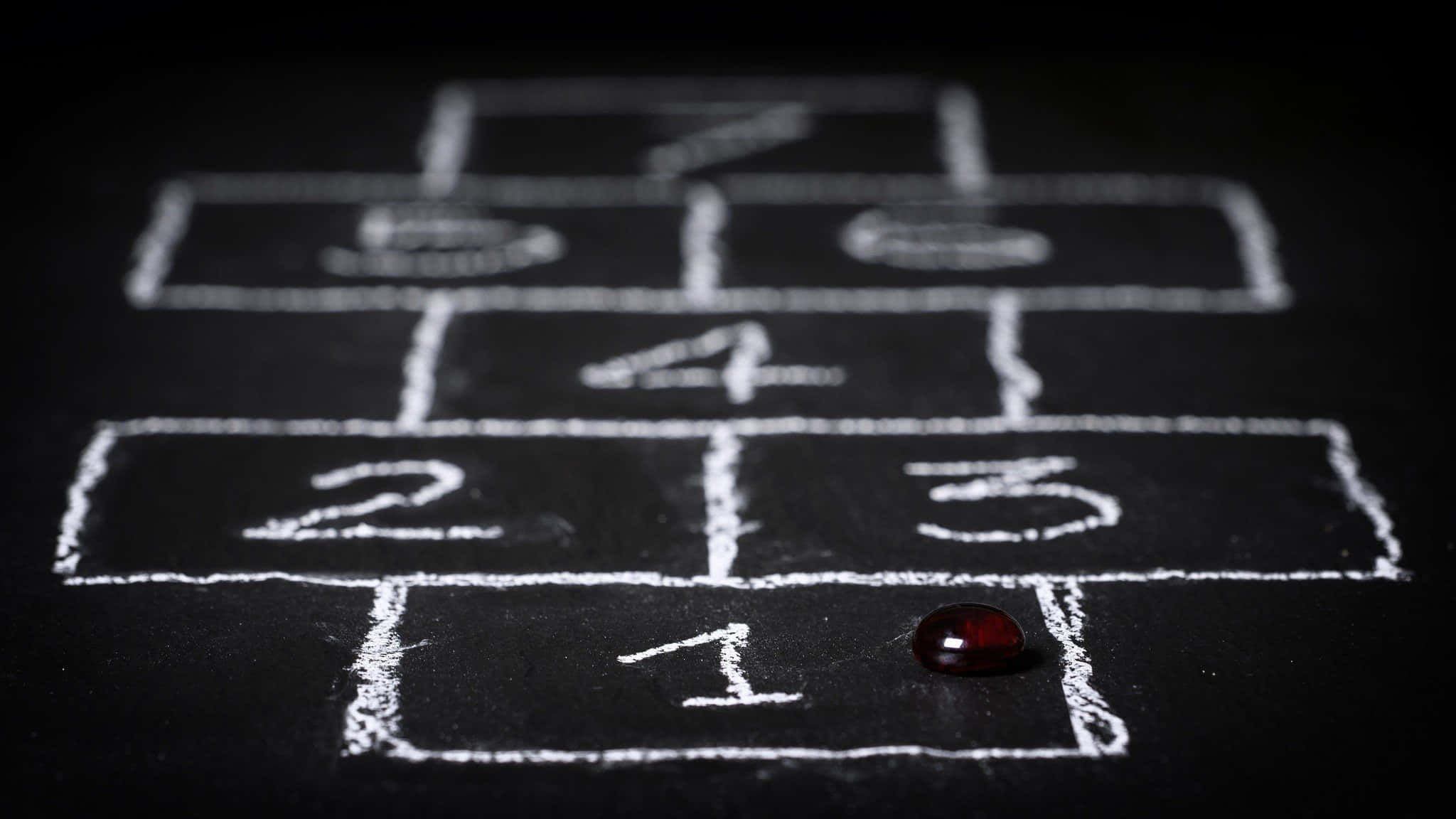 Educational Games 桌布 壁紙 — 一個用粉筆喺深色表面上畫嘅跳飛機格仔嘅特寫，一粒紅色波子喺第一格上面。