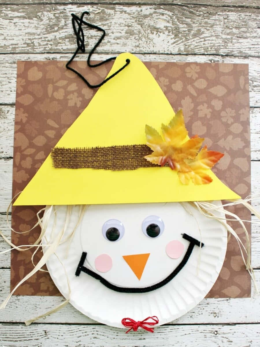 Fall Crafts 桌布 壁紙 — 一個用紙碟製作、充滿歡樂的稻草人手工艺，配有黃色帽子、秋葉和稻草頭髮，以鄉村木質背景襯托。