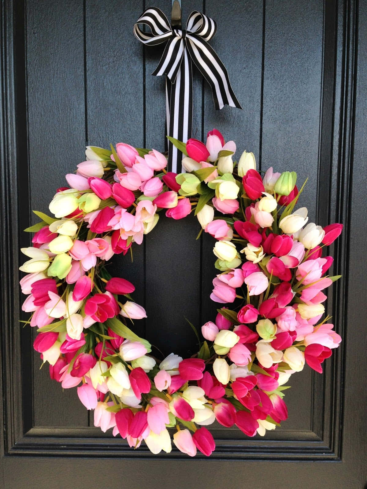 Spring Wreaths 桌布 壁紙 — 一個由粉紅、白色和紅色鬱金香組成的鮮豔花環掛在深色木門上，門上裝飾著黑白條紋蝴蝶結。