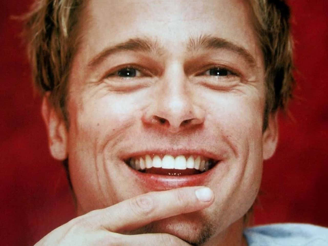 Brad Pitt 桌布 壁紙 — 一個男人嘅特寫肖像，佢帶住溫暖嘅笑容，手放喺下巴附近，背景係鮮艷嘅紅色。