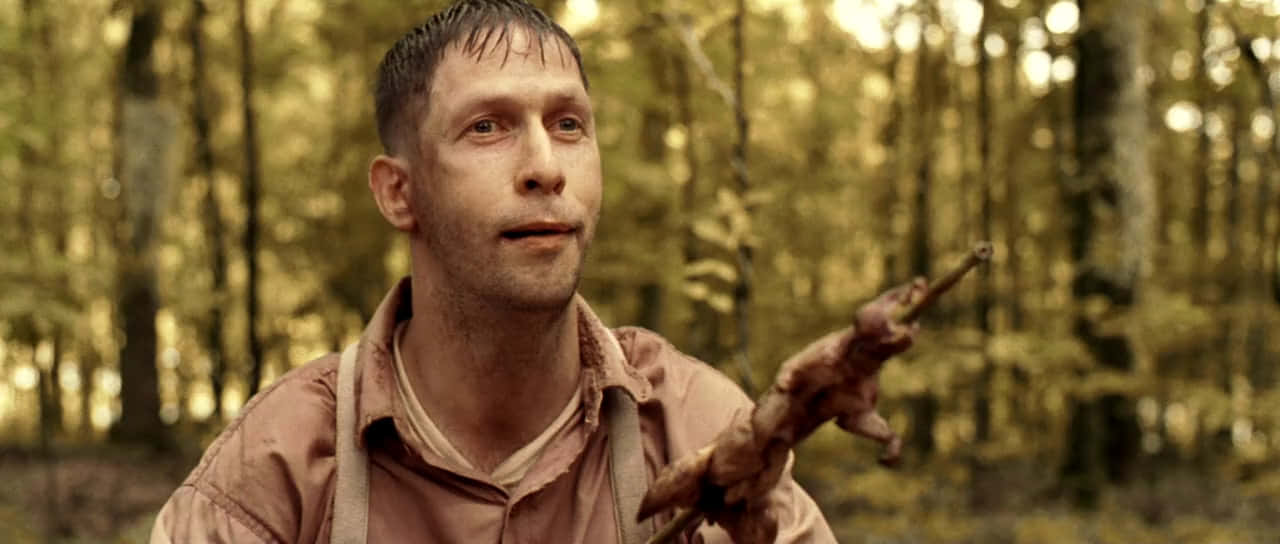 Tim Blake Nelson 桌布 壁紙 — 一個男人在森林裏拿着烤好的動物放在火上，表情驚訝或擔憂。