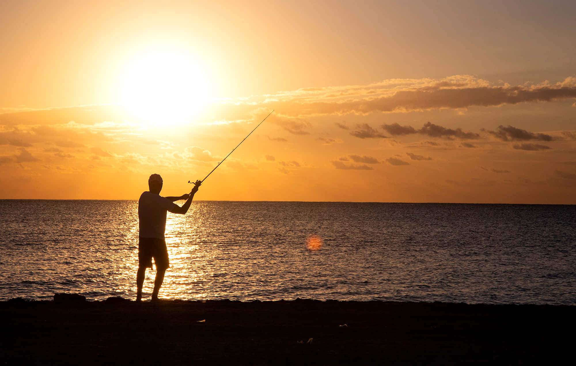 Fishing Rod 桌布 壁紙 — 一個男人在金色日落、雲彩和閃爍海水中拋出魚竿的剪影。