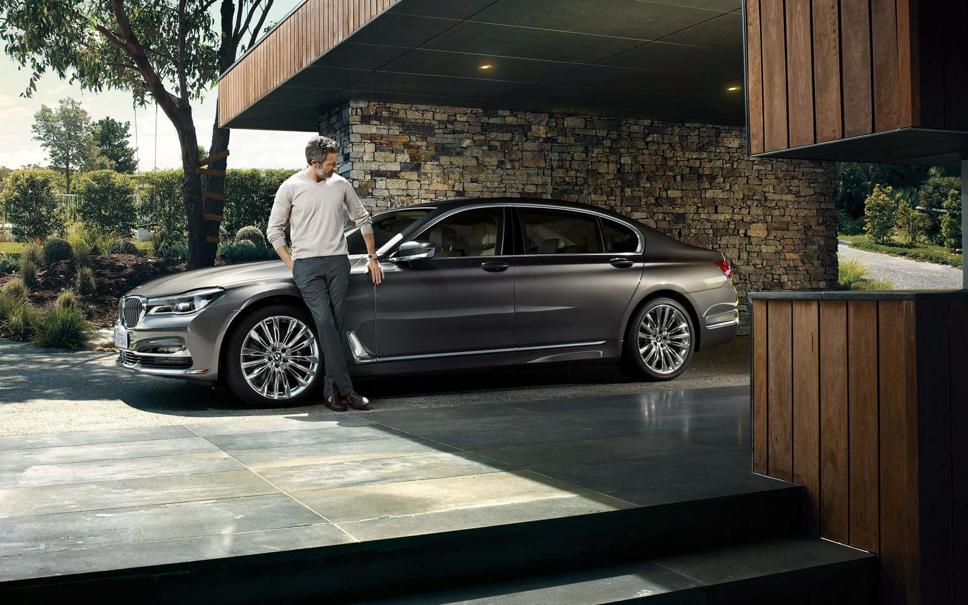Bmw 7 Series 桌布 壁紙 — 一個男人在陽光明媚的日子裡，隨意靠在一輛停放在現代石木車棚下的時尚灰色豪華轎車上。