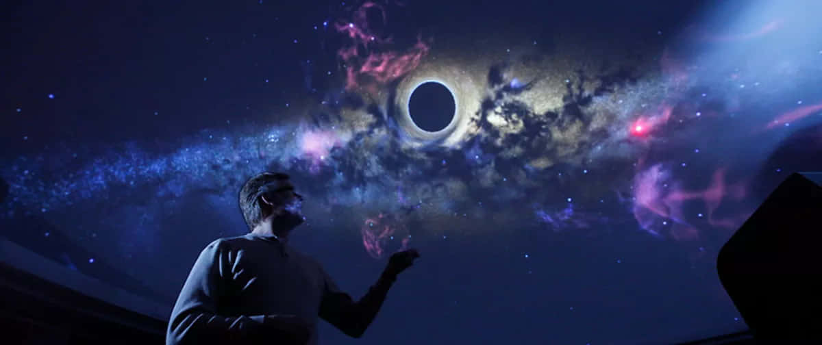 Planetarium 桌布 壁紙 — 一個男人在黑暗的房間裏抬頭凝視著一個令人驚嘆的宇宙投影，其中包含一個黑洞和充滿活力的星雲。