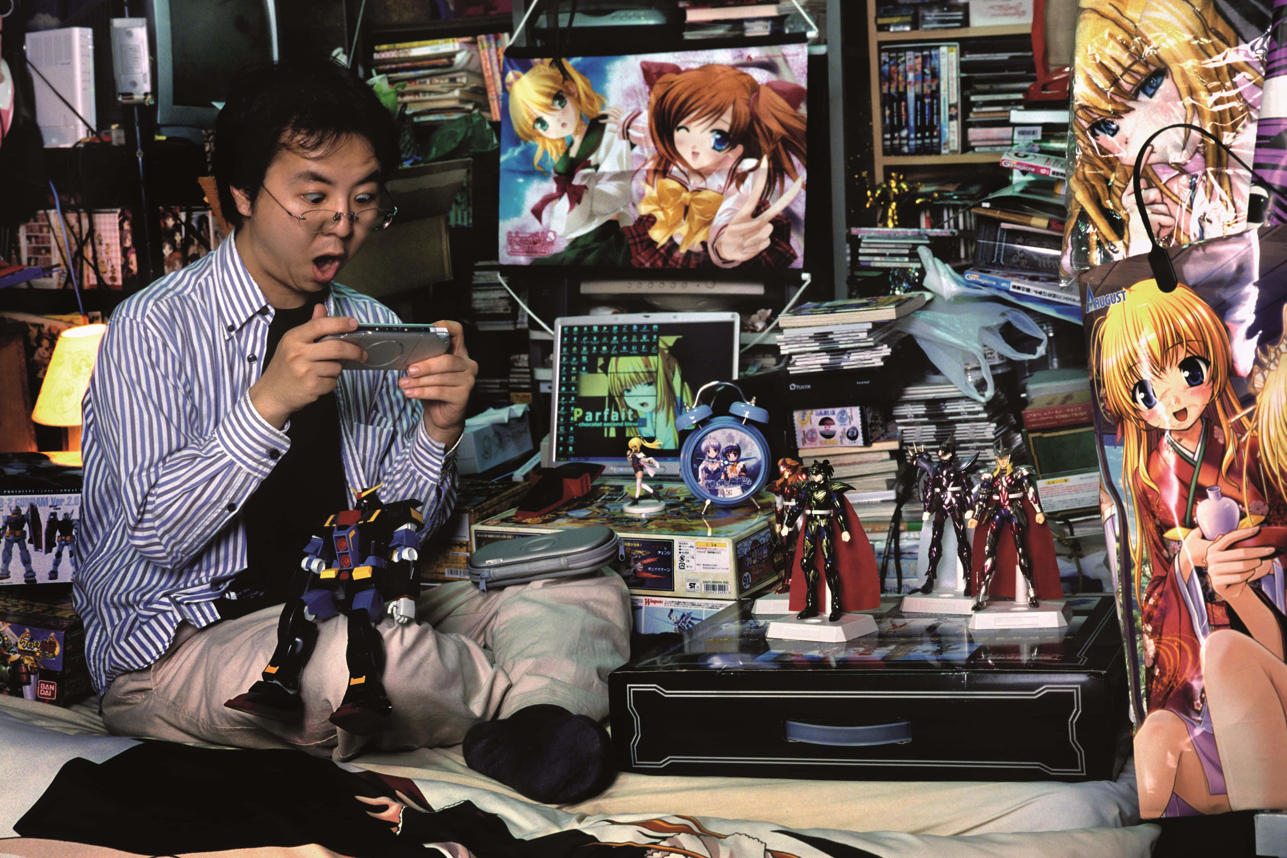 Otaku 桌布 壁紙 — 一個男人坐在床上，周圍是動漫海報、人偶和遊戲裝備，專注於他的手持設備。