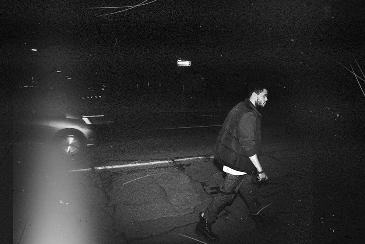  壁紙 — 一個男人夜晚獨自走在裂開嘅行人路上，旁邊有架泊咗嘅車，喺昏暗嘅街燈下，黑白畫面。
