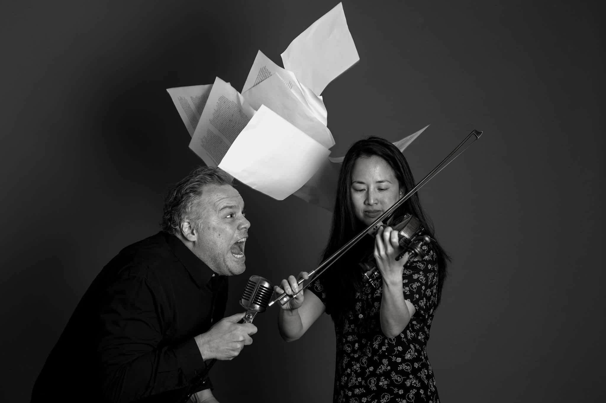 Vincent D'onofrio 桌布 壁紙 — 一個男人對著復古咪高峰尖叫，紙張在一個拉小提琴的女人上方爆炸，場景在鮮明的黑白攝影棚中。