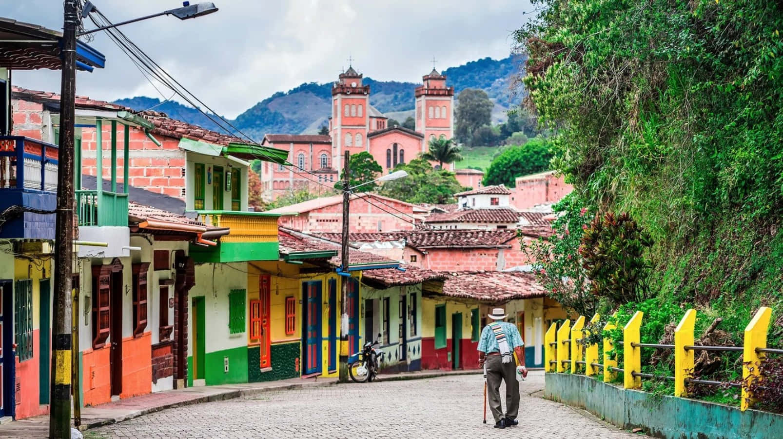Colombia 桌布 壁紙 — 一個男人沿著一條鵝卵石街道走下來，兩旁是色彩繽紛的殖民時期房屋和一座綠色山丘上的教堂。