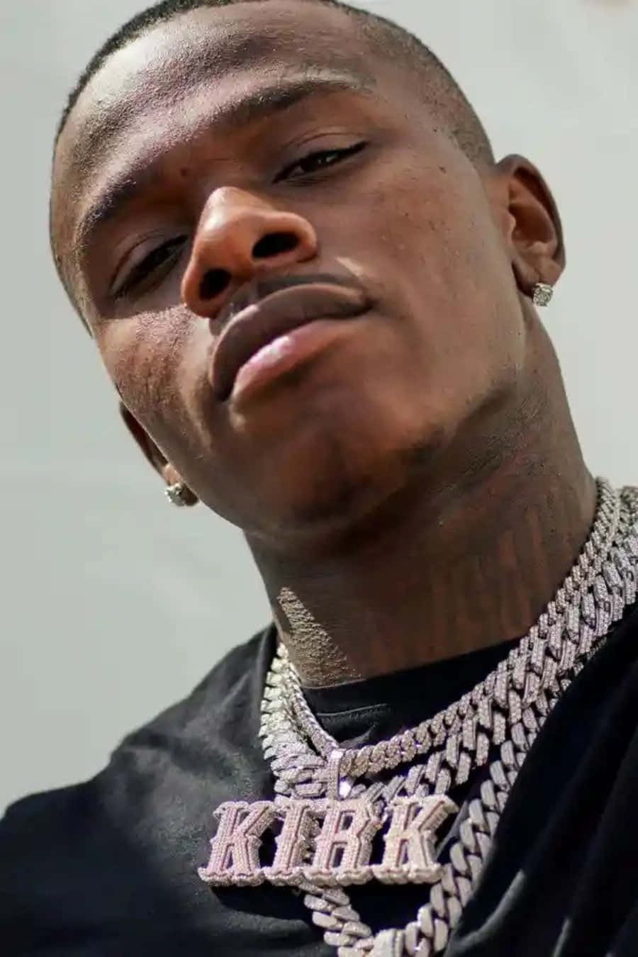 Dababy 桌布 壁紙 — 一個男人的特寫肖像，戴著鑲有鑽石的鏈條，黑色襯衫上有 'KIRK' 字樣，背景簡單。