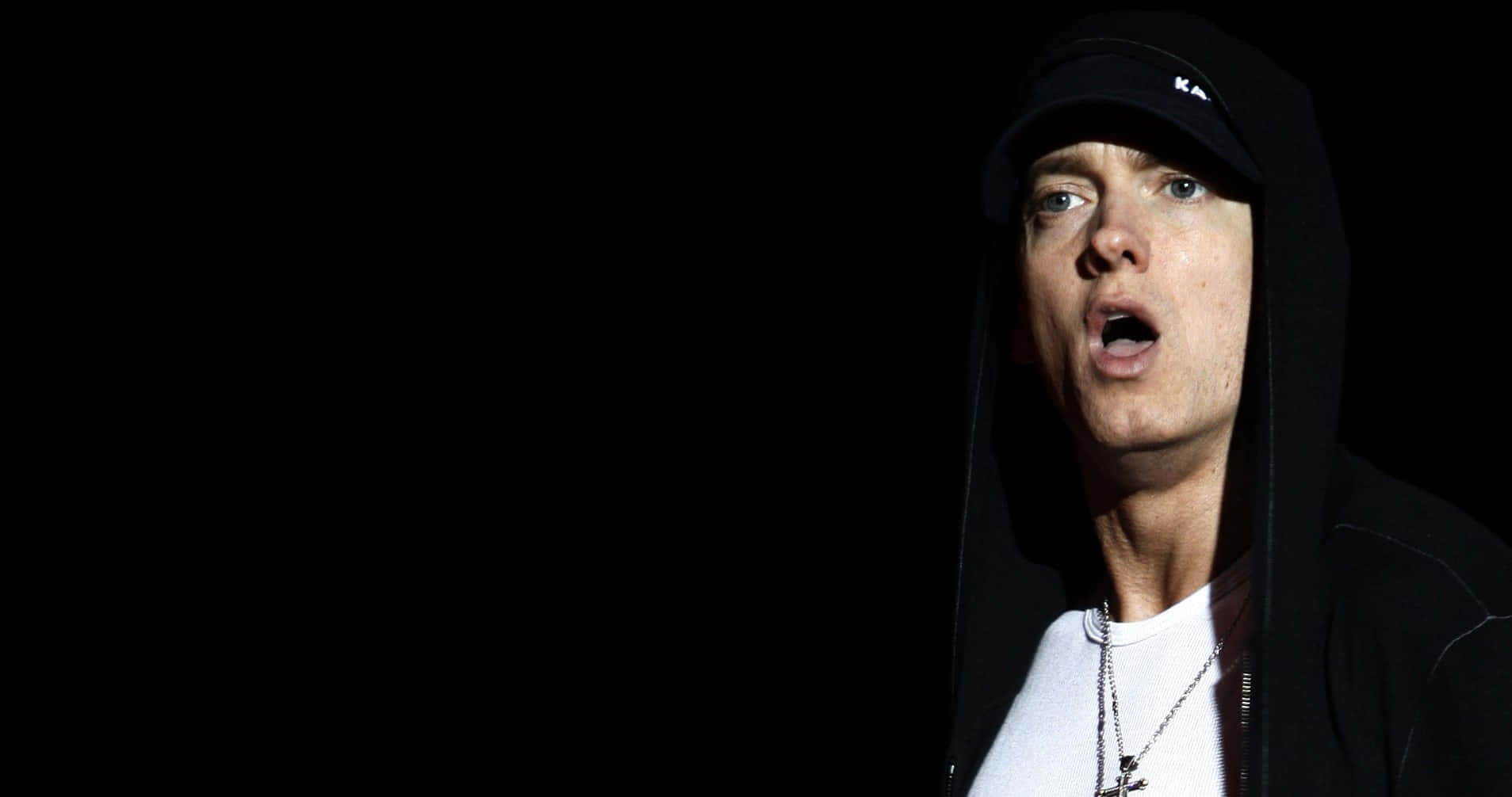 Eminem 桌布 壁紙 — 一個男人穿著黑色連帽衫和帽子，在強烈燈光下以純黑背景拍攝的戲劇性特寫。