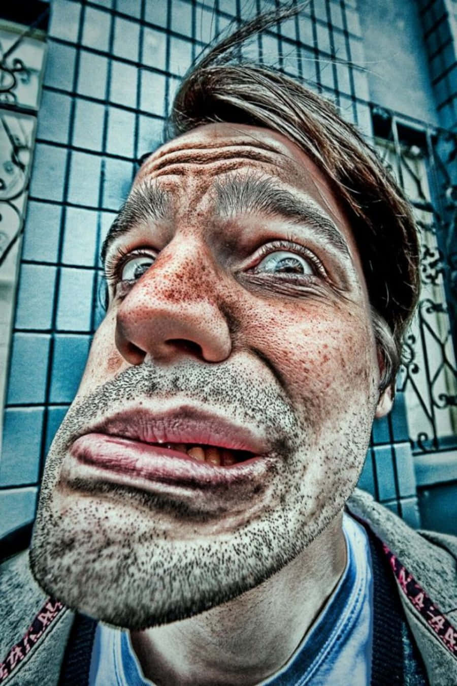 Funny Faces 桌布 壁紙 — 一個男人臉部的扭曲特寫，特徵誇張，眼睛睜大，背景是瓷磚牆。