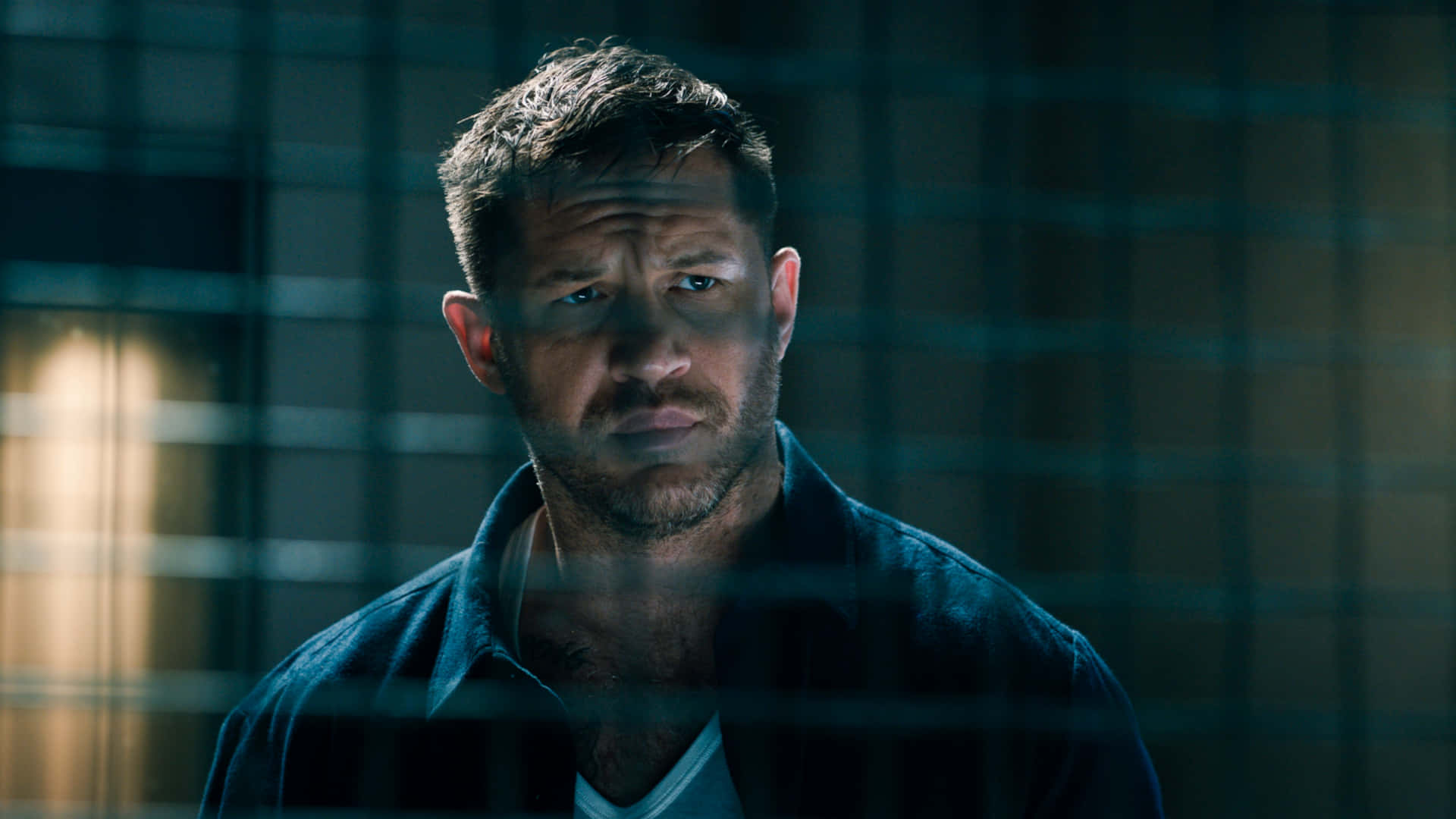 Tom Hardy Venom 桌布 壁紙 — 一個留有鬍渣的男人在昏暗的藍色燈光下，背後有溫暖的光源，專注地透過監獄鐵欄杆凝視。