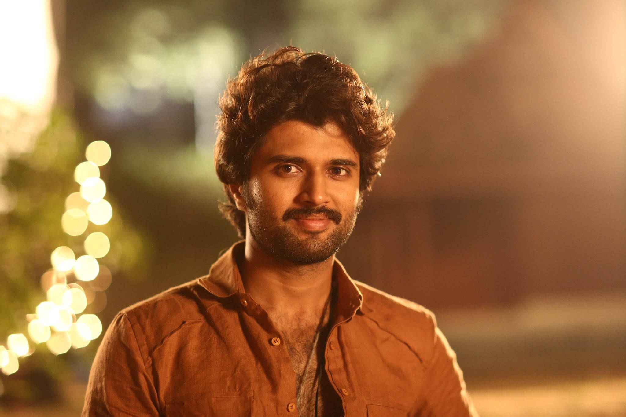 vijay devarakonda 4K 桌布 壁紙 — 一個留著捲髮和鬍鬚的男人，在溫暖的散景夜間背景中溫柔地微笑。