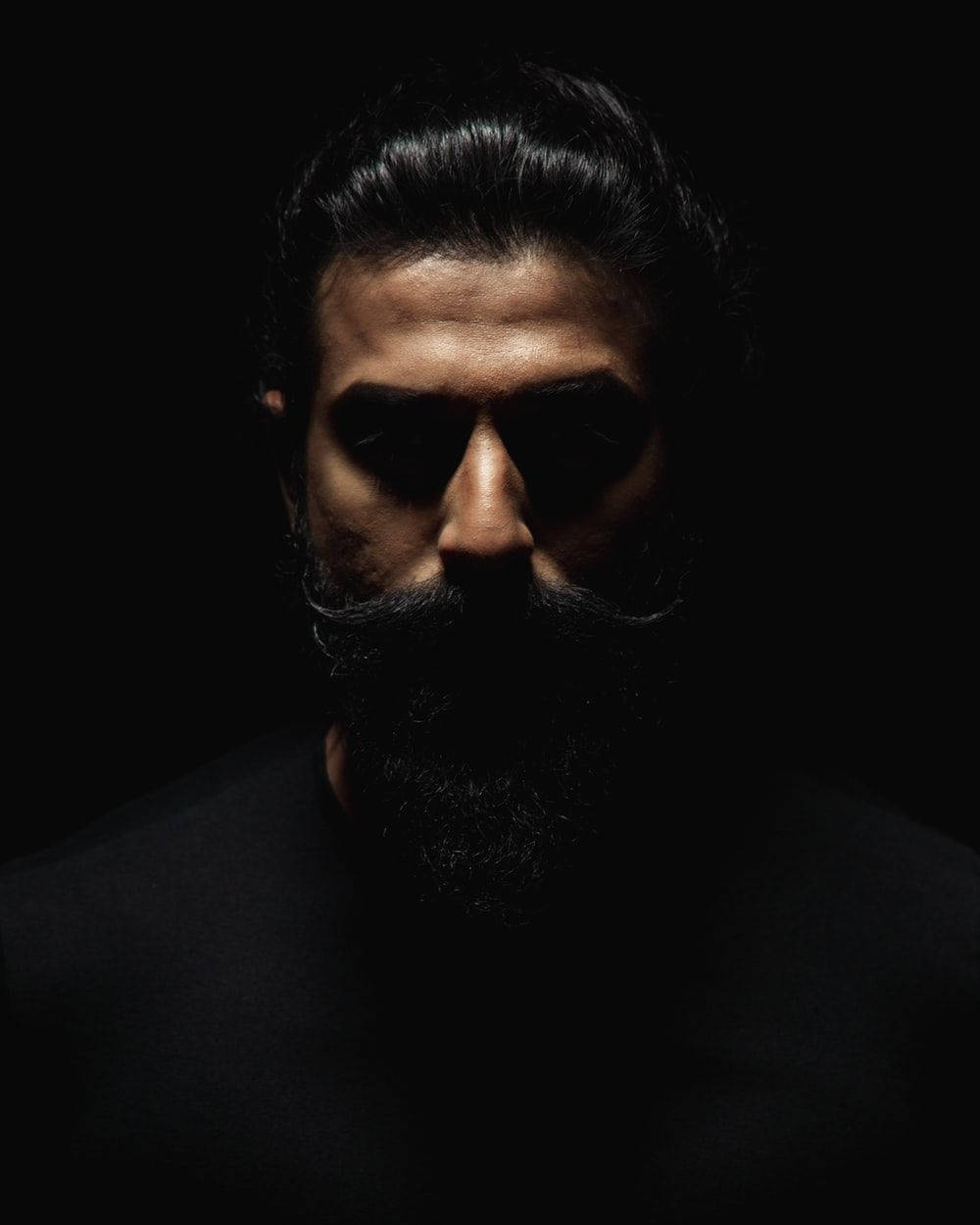 beard 桌布 壁紙 — 一個留著濃密鬍鬚、頭髮向後梳的男人，在近乎黑暗的環境中，在戲劇性的側光下，眼神銳利地凝視著。