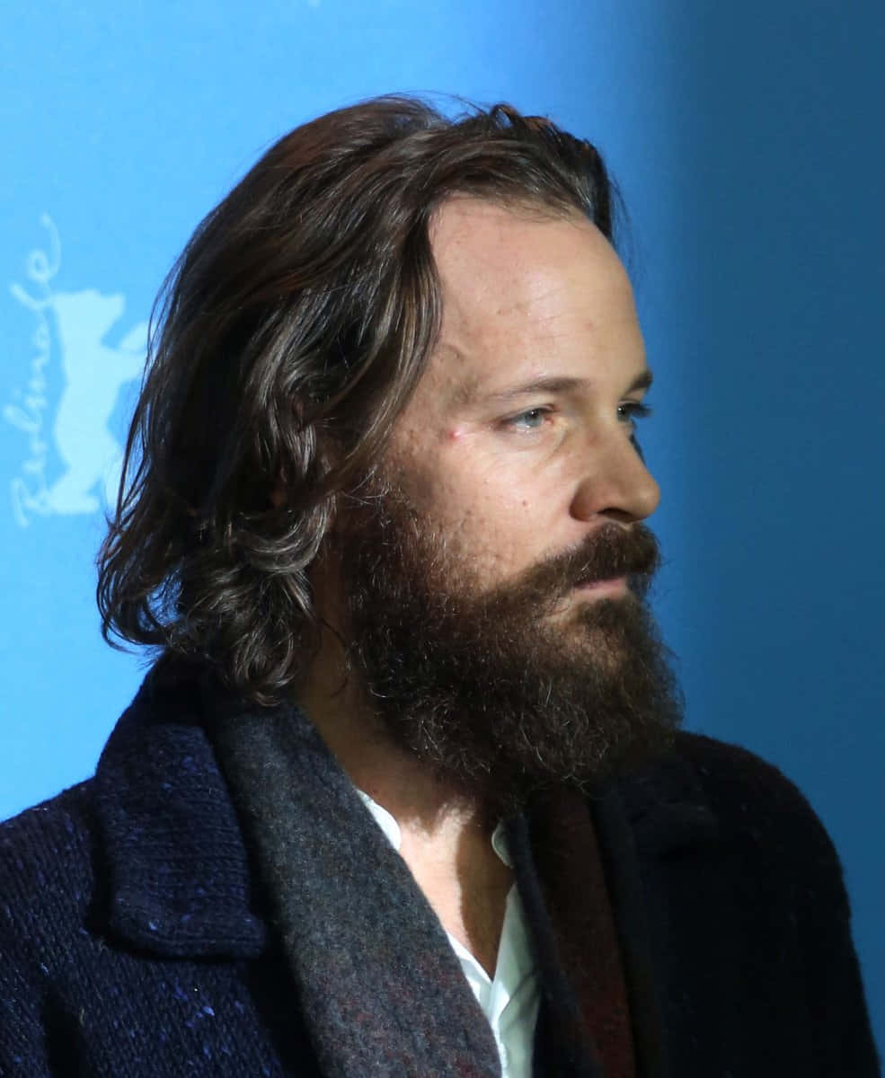 Peter Sarsgaard 桌布 壁紙 — 一個留著濃密鬍鬚和波浪頭髮的男人，在帶有淡色標誌的純藍色背景旁望向一邊。