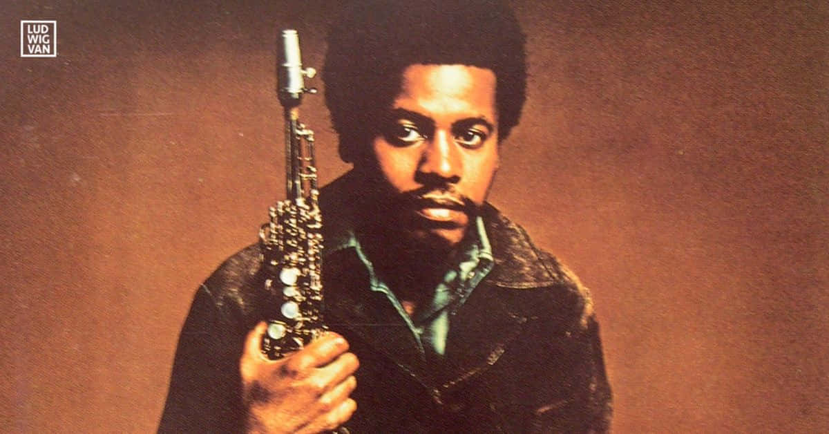Wayne Shorter 桌布 壁紙 — 一個留著爆炸頭的男人在溫暖的棕色背景前拿著薩克斯風的復古工作室肖像。